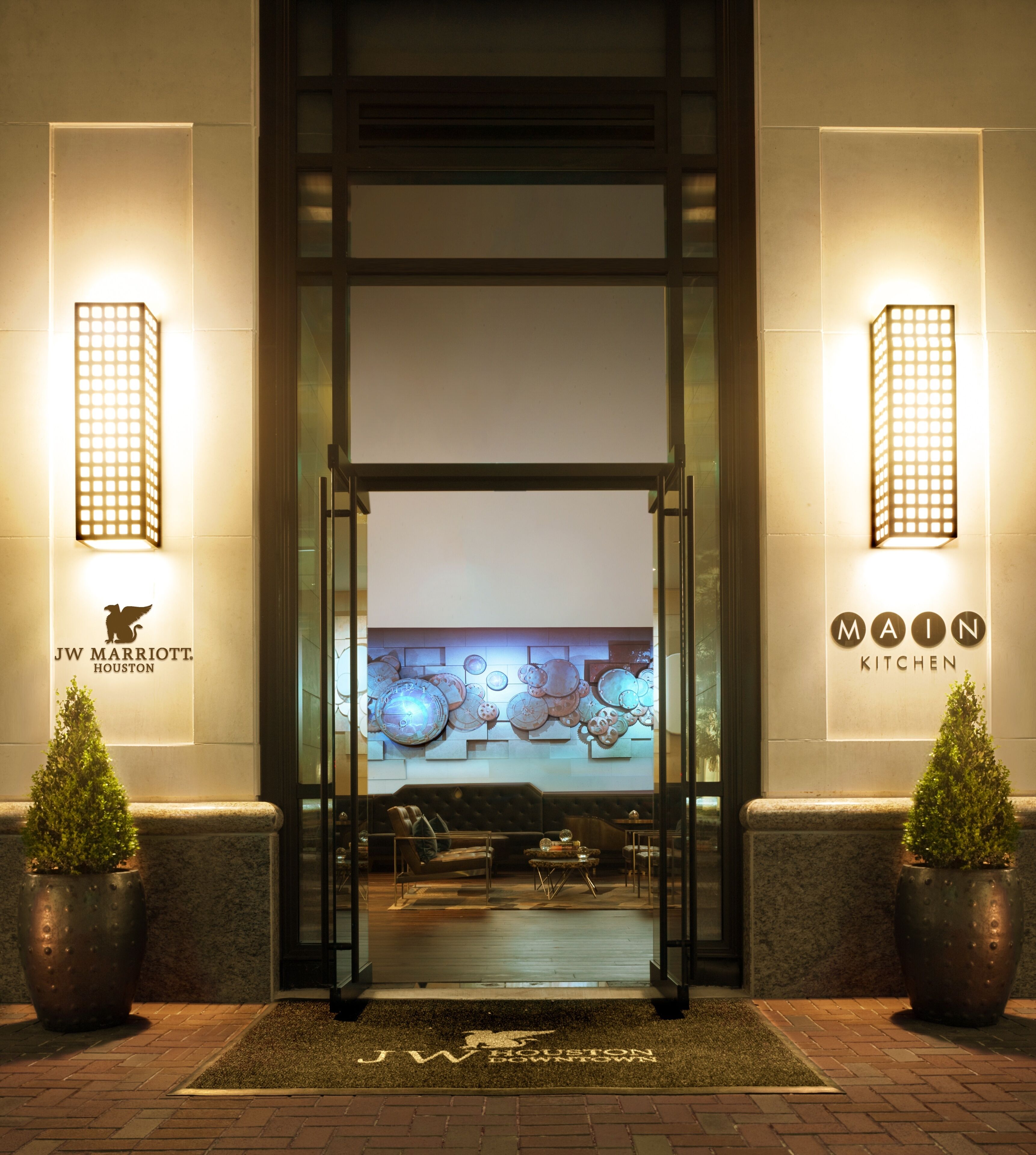 L’entrée principale du JW Marriott Houston présente un design moderne, avec une porte en verre, un espace d’accueil doté d’un canapé et de fauteuils, et une œuvre d’art décorative au-dessus de l’entrée.