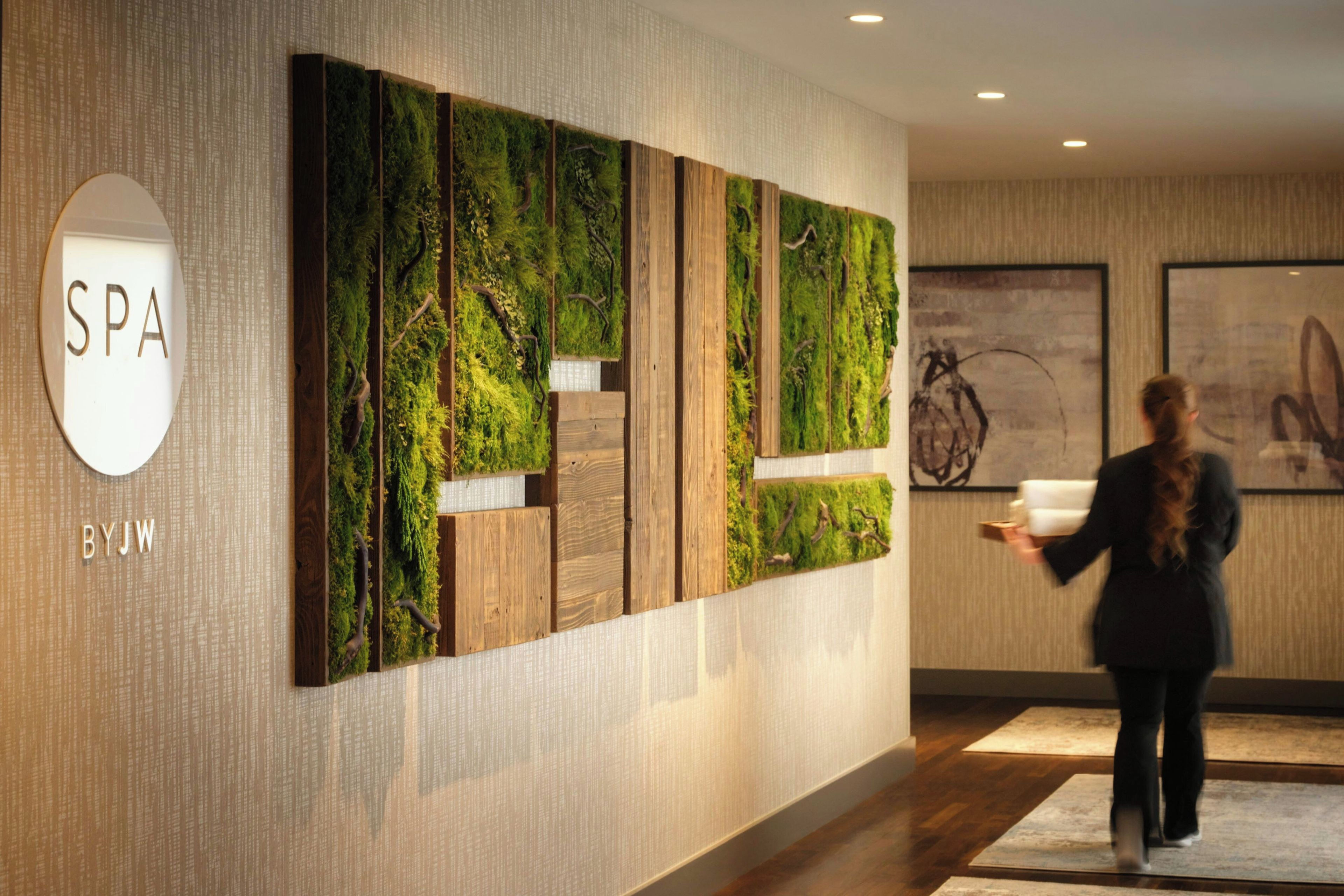 A spa sign with 'SPA' and 'BYJW' on a wall, a large vertical wooden panel with a green moss-like texture, and a person carrying a tray.