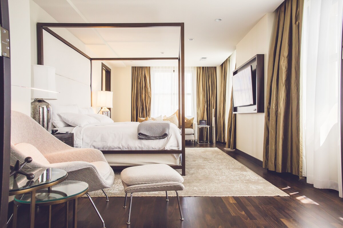 A modern hotel room with a four-poster bed, a glass coffee table, a cushioned bench, and a flat-screen TV.