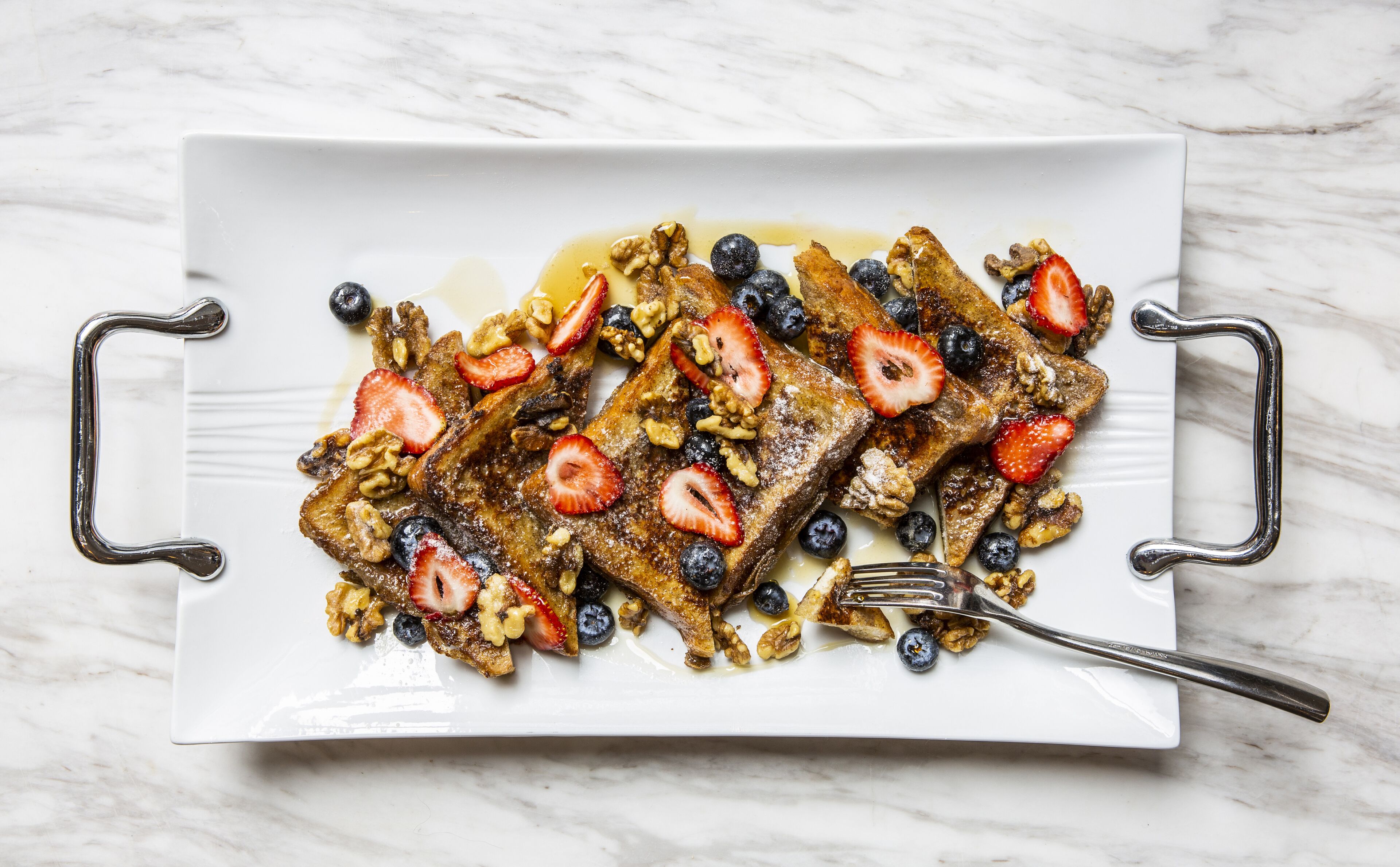 A rectangular white plate with a handle on each side, containing French toast topped with blueberries, strawberries, and walnuts, and drizzled with syrup.