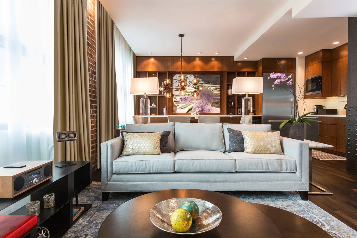 A modern living room with a grey sofa, a coffee table with decorative items, a chandelier, and a kitchen area in the background.