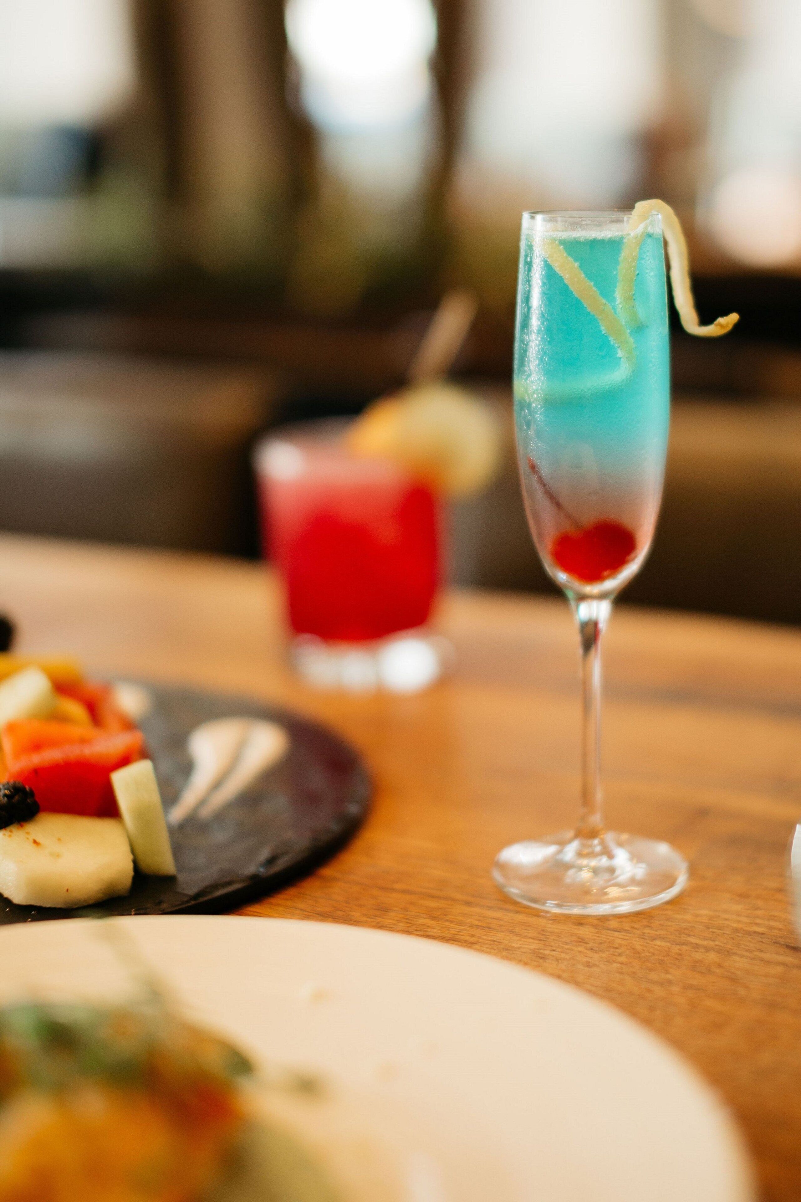 A table with a glass of blue cocktail garnished with a lemon twist, a plate with various food items, and a red drink in the background.