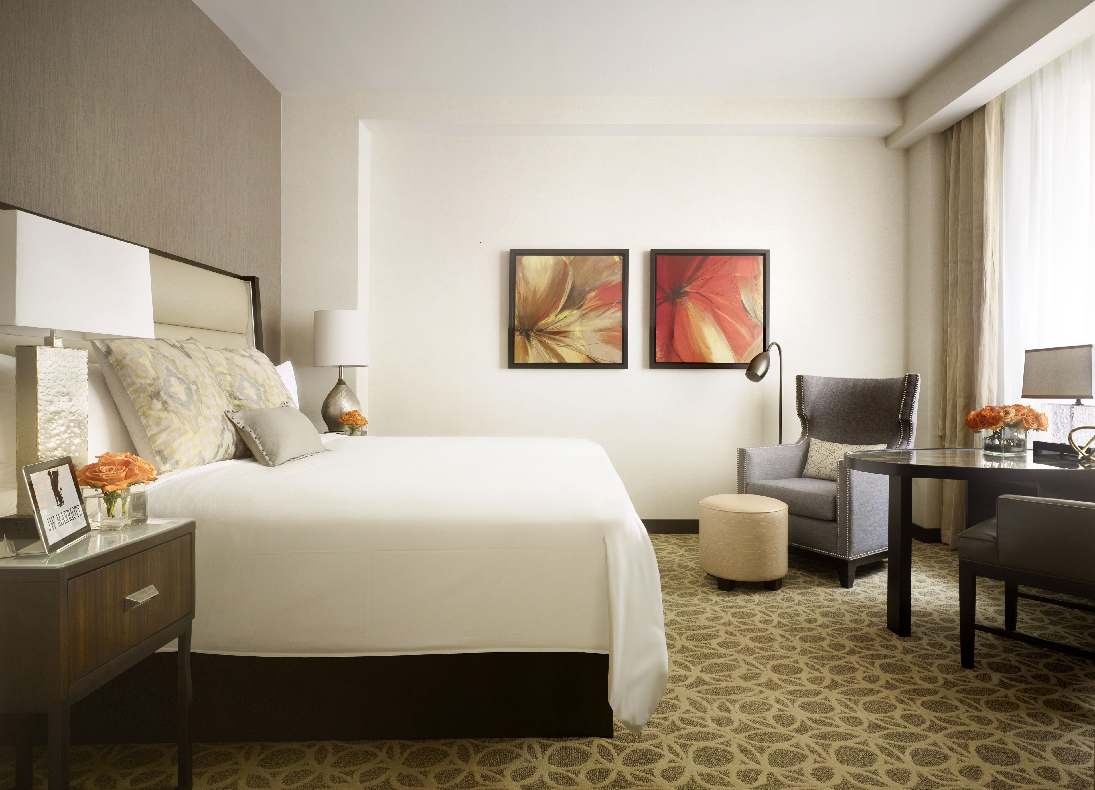 A hotel room with a large bed, a desk, a chair, a lamp, and two framed floral paintings on the wall.