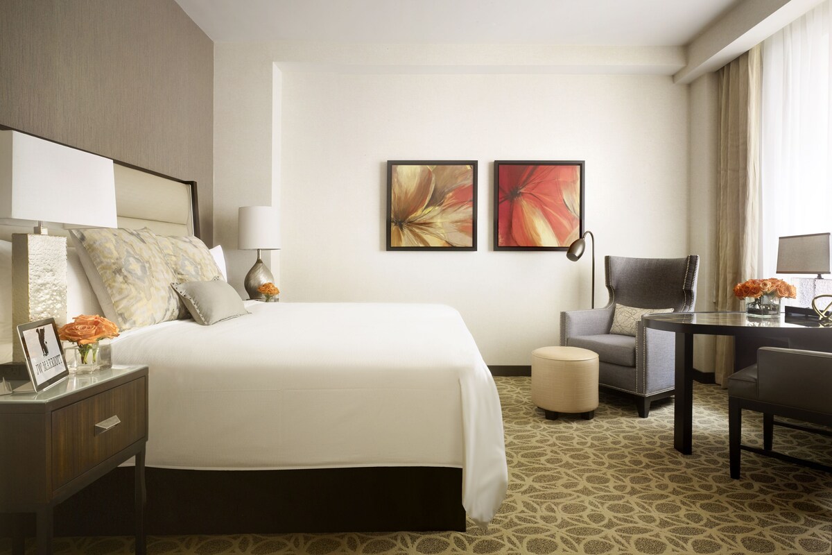 A hotel room with a large bed, a desk, a chair, a lamp, and two framed floral paintings on the wall.