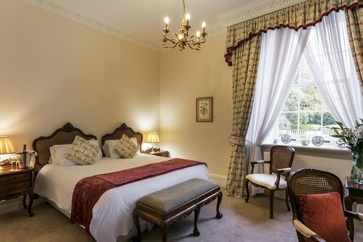 A bedroom with a large bed, two bedside tables with lamps, a chair, a window with curtains, and a chandelier.