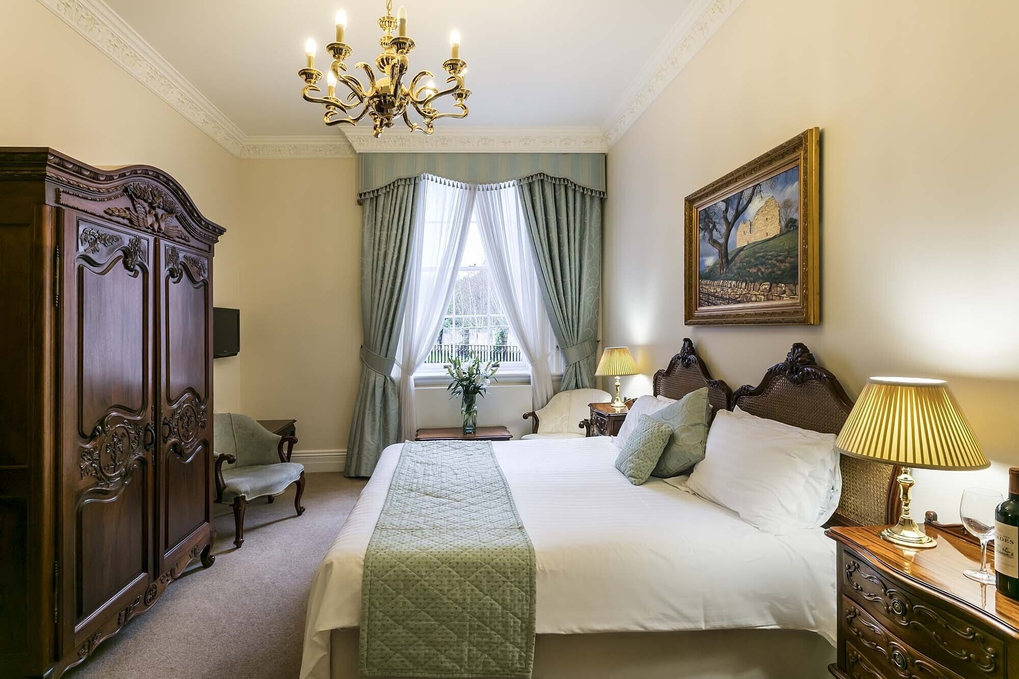 A bedroom with a large bed, a wooden wardrobe, a chandelier, a window with curtains, and a painting on the wall.