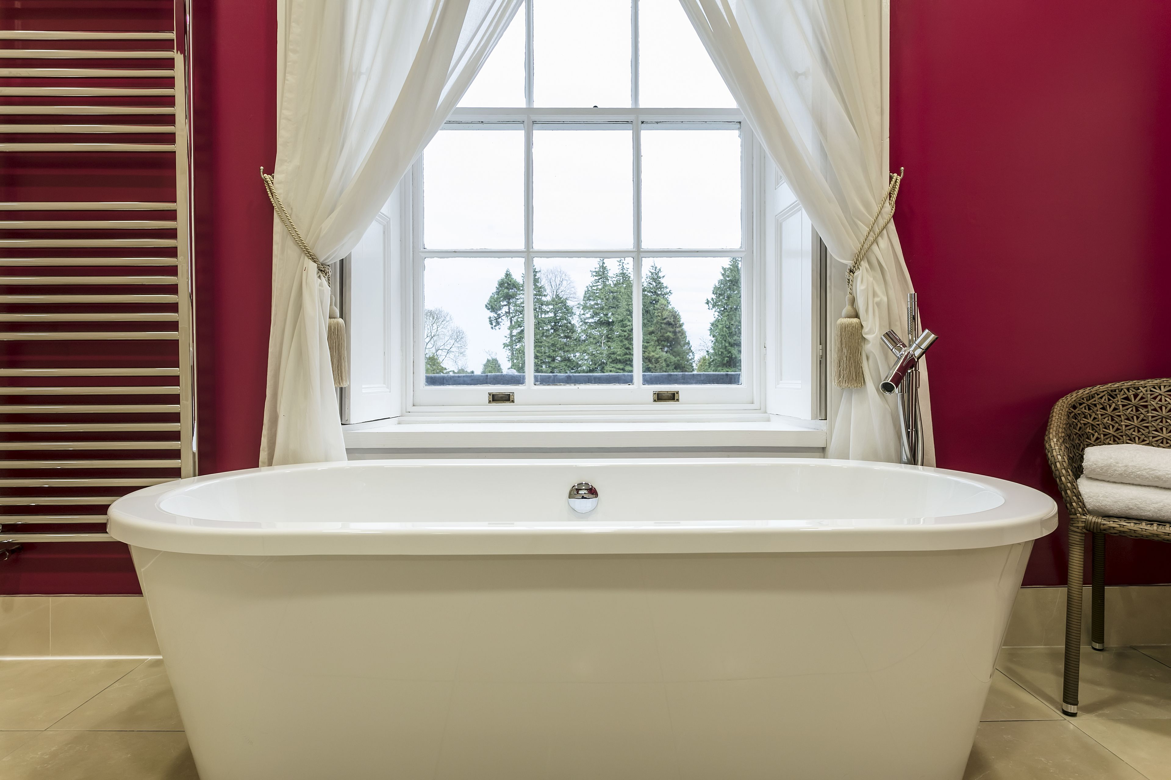 A bathroom with a freestanding bathtub, a window with curtains, and a wicker chair with a towel.