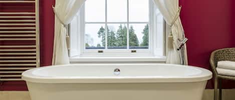 A bathroom with a freestanding bathtub, a window with curtains, and a wicker chair with a towel.