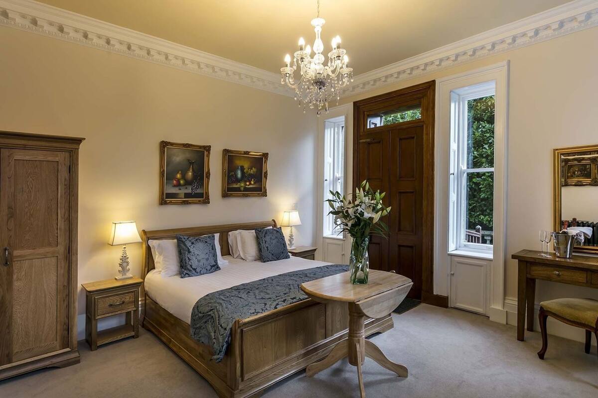 A bedroom with a wooden bed, two bedside tables with lamps, a chandelier, a wooden desk with a chair, and a window with a view of greenery.