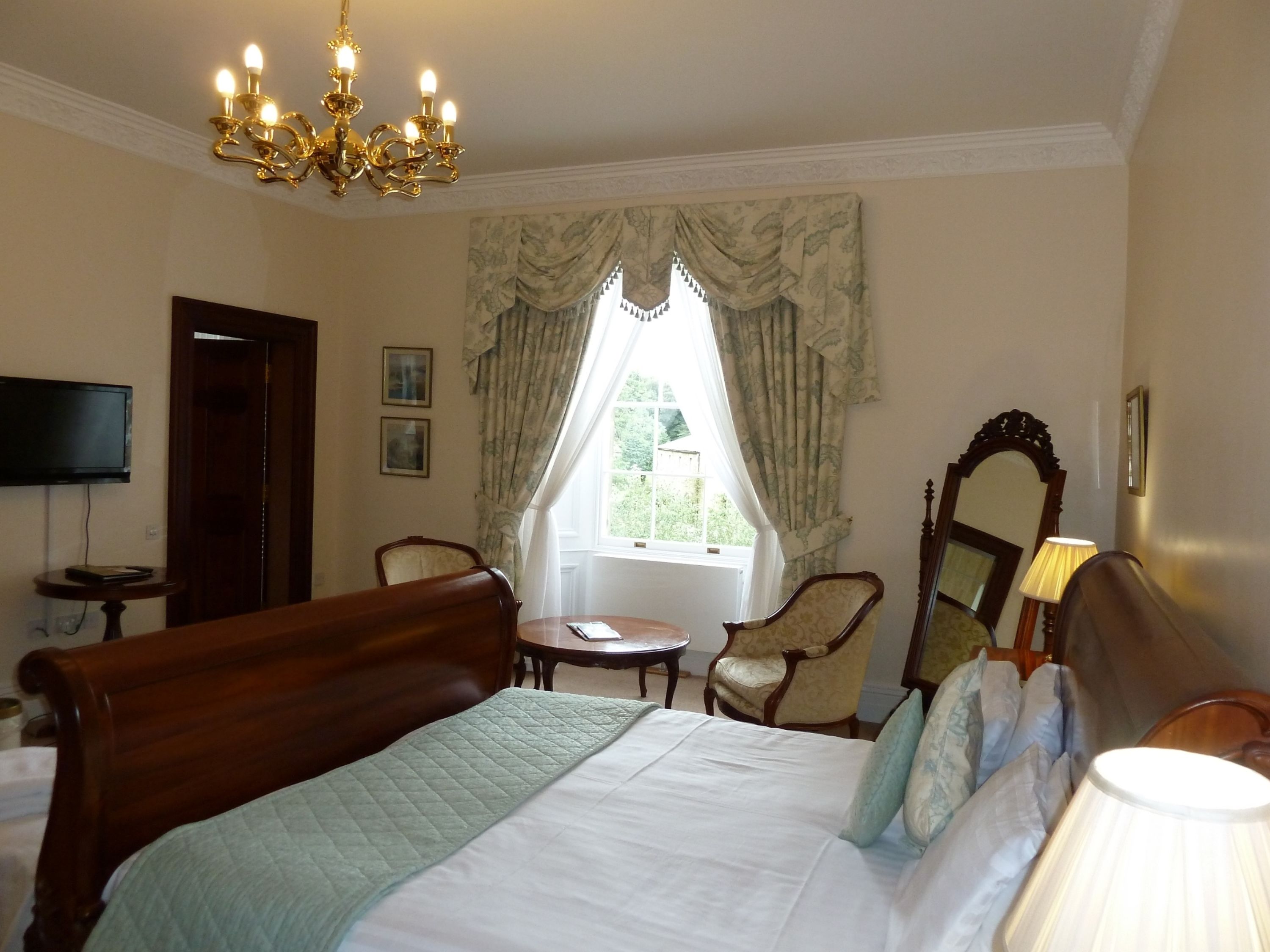 A hotel room with a large bed, a chandelier, a television, a window with curtains, a mirror, and a lamp.