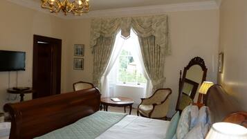 A hotel room with a large bed, a chandelier, a television, a window with curtains, a mirror, and a lamp.