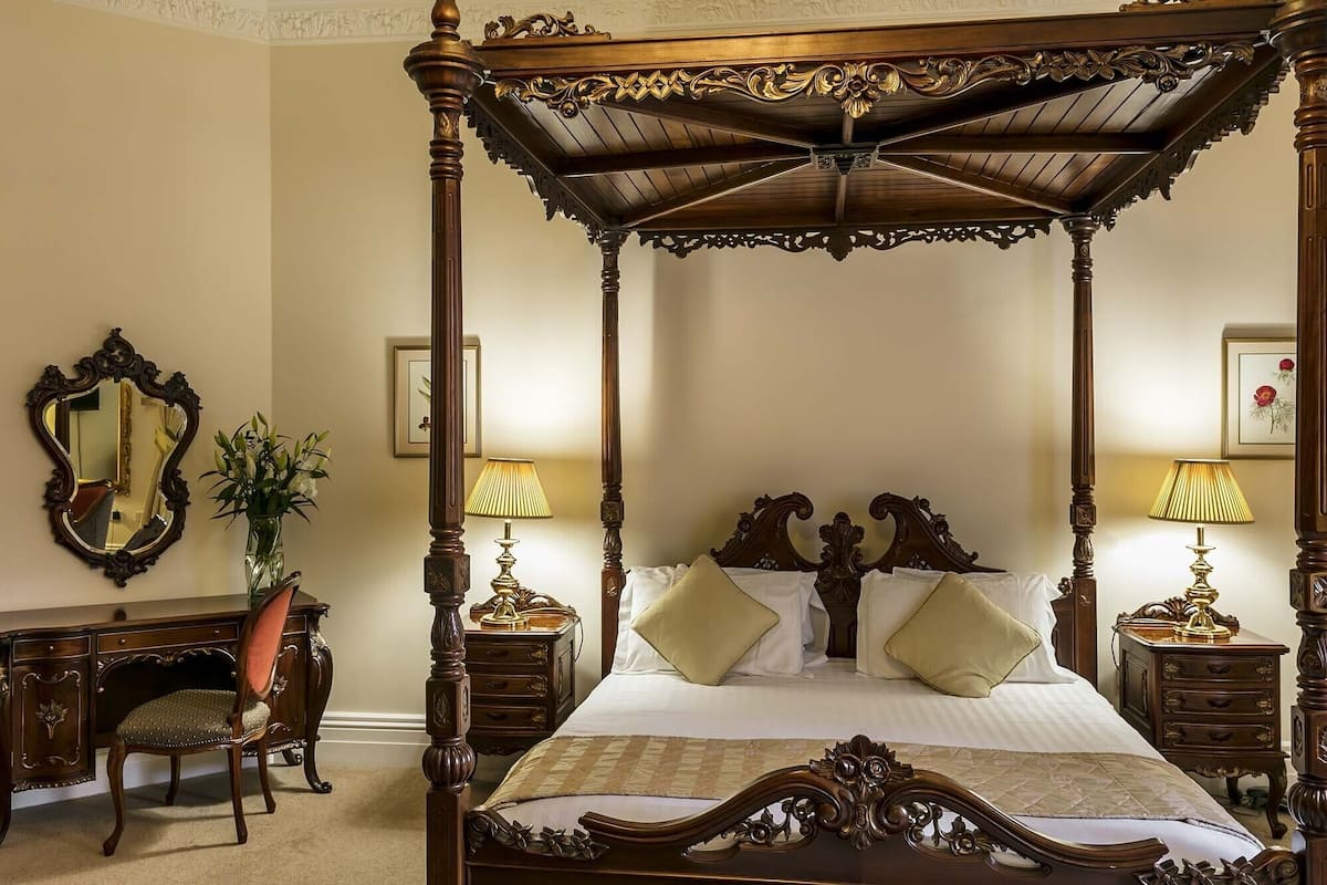 A four-poster canopy bed with ornate wooden carvings, two bedside tables with lamps, a chair, and a desk with a mirror and vase.