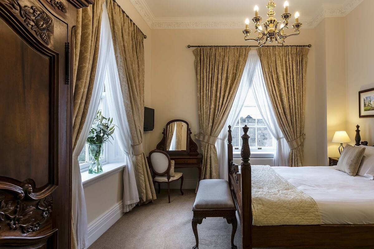 A bedroom with a large bed, a chair, a desk, a mirror, and a window with curtains.