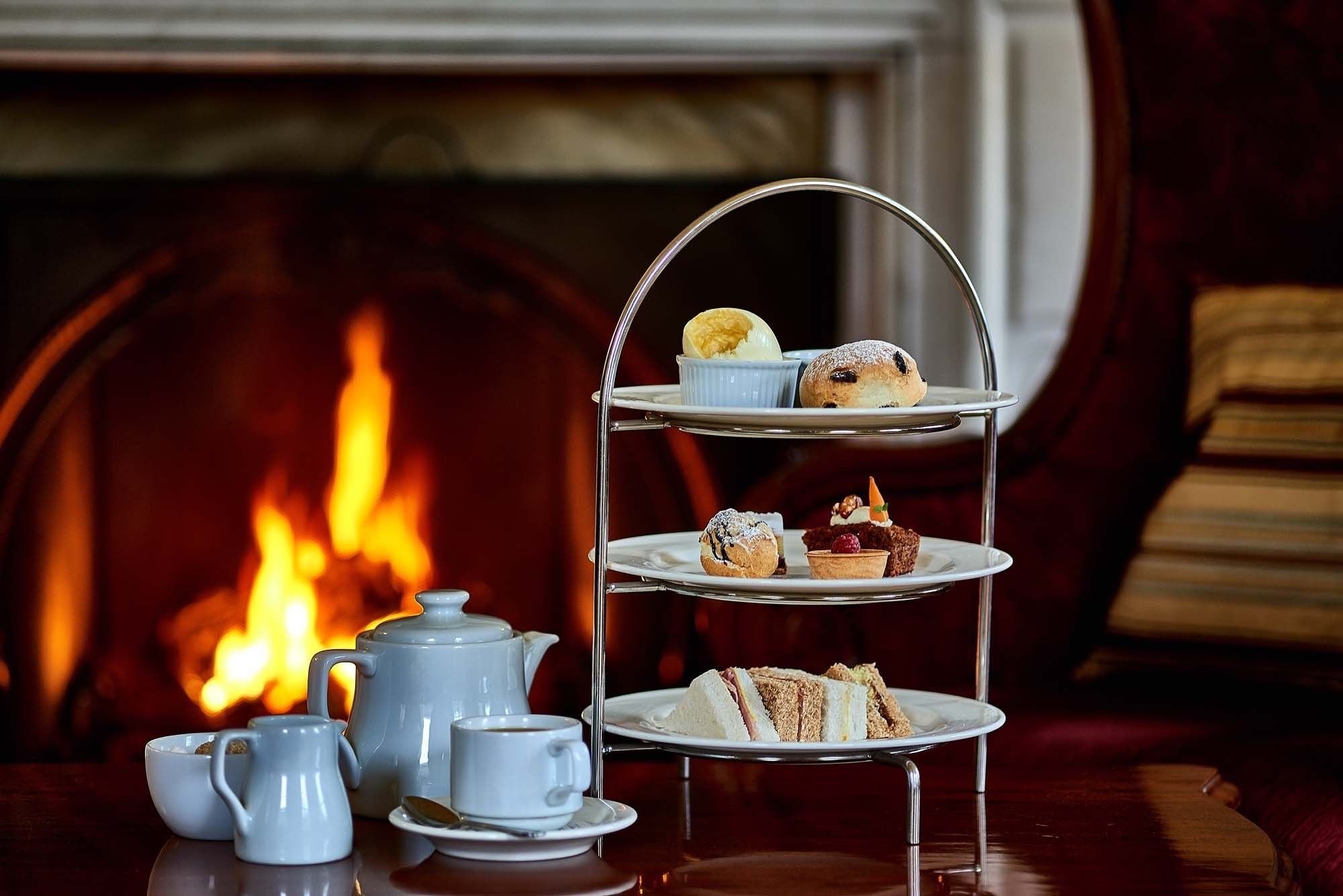 Un soporte de tres niveles para postres con diversos pasteles y una tetera sobre una mesa frente a una chimenea.