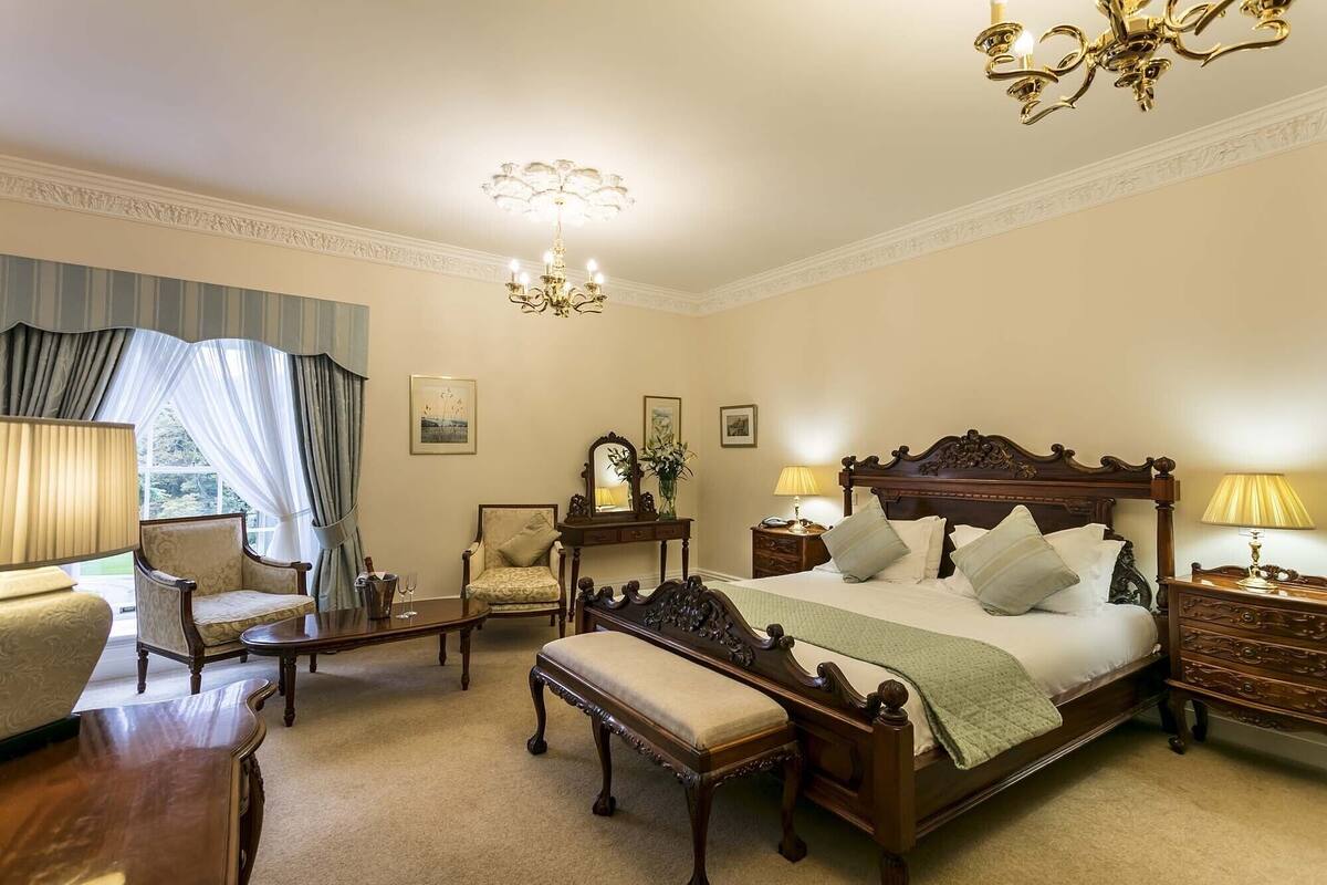 A classic, elegantly furnished bedroom with a large bed, a sitting area with a chair and table, and a chandelier.