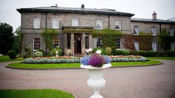 A large stone building with a formal entrance, a well-maintained garden with flower beds, and a gravel driveway.