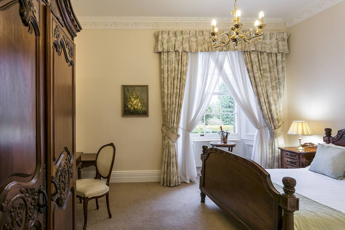 A bedroom with a wooden bed, a chair, a nightstand with a lamp, and a window with curtains.