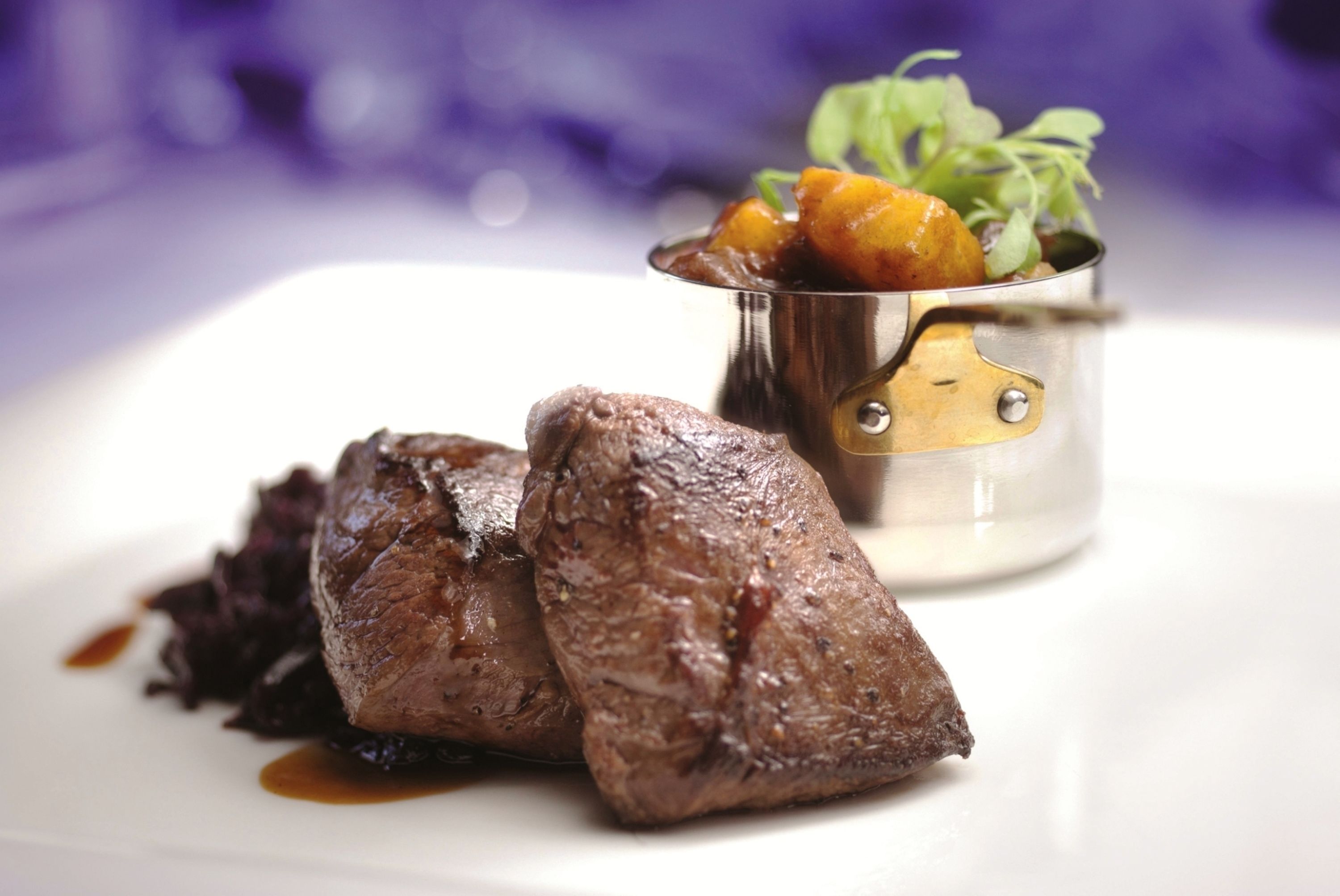 A plate with two pieces of meat and a small metal cup with vegetables.