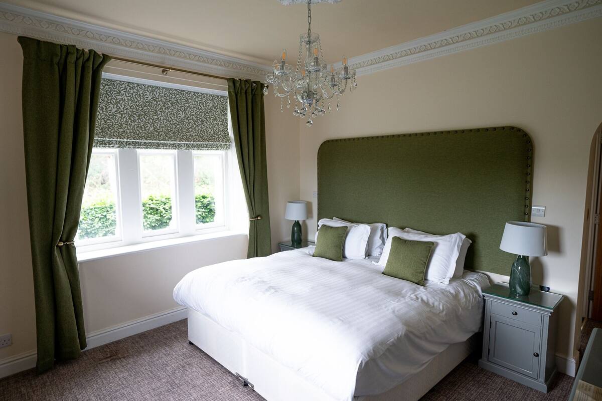A neatly arranged bedroom with a large bed, green curtains, a chandelier, and two bedside tables with lamps.