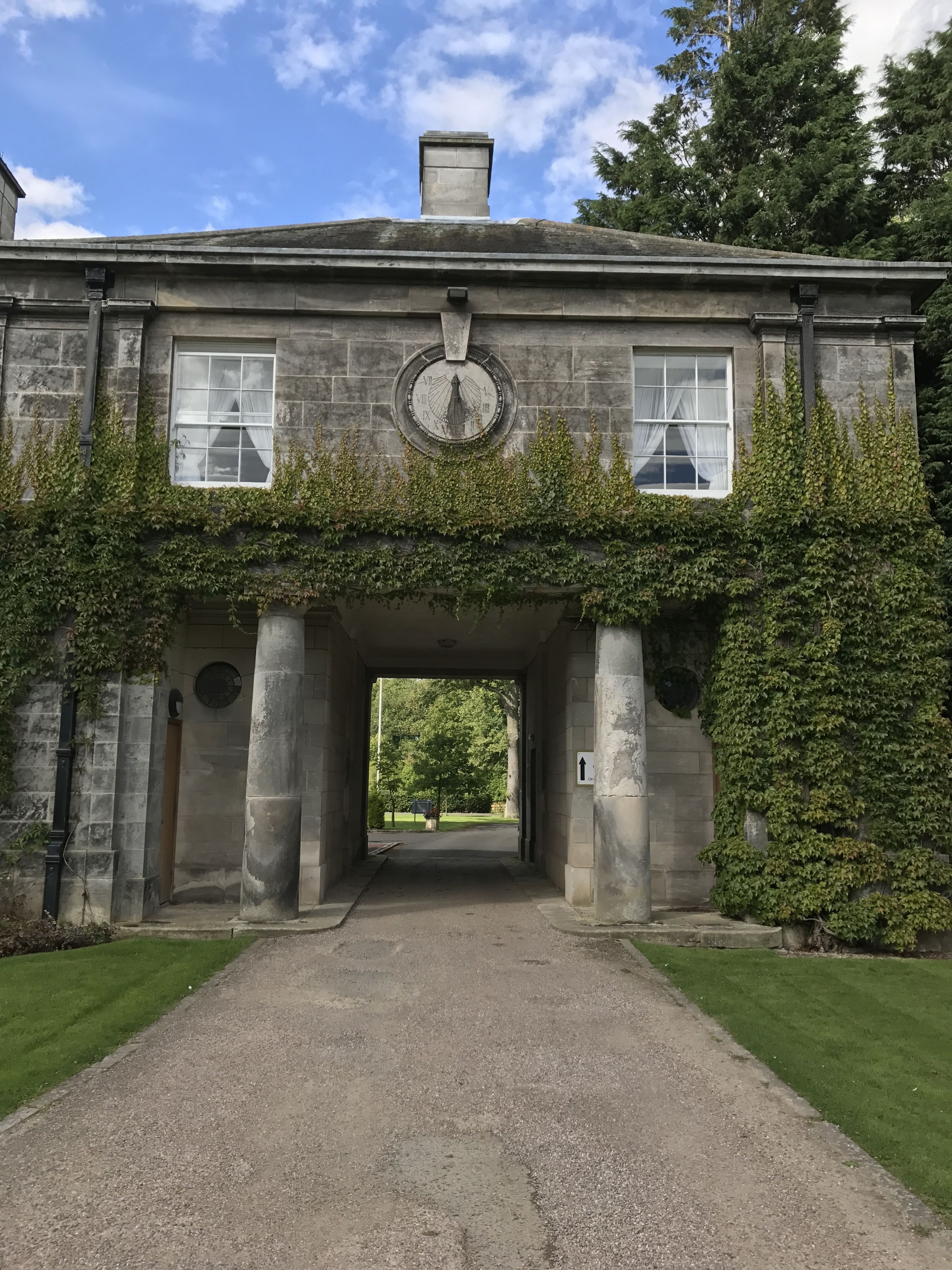 A stone building with a central archway, a sundial, and a grassy pathway leading to a tree-lined area.