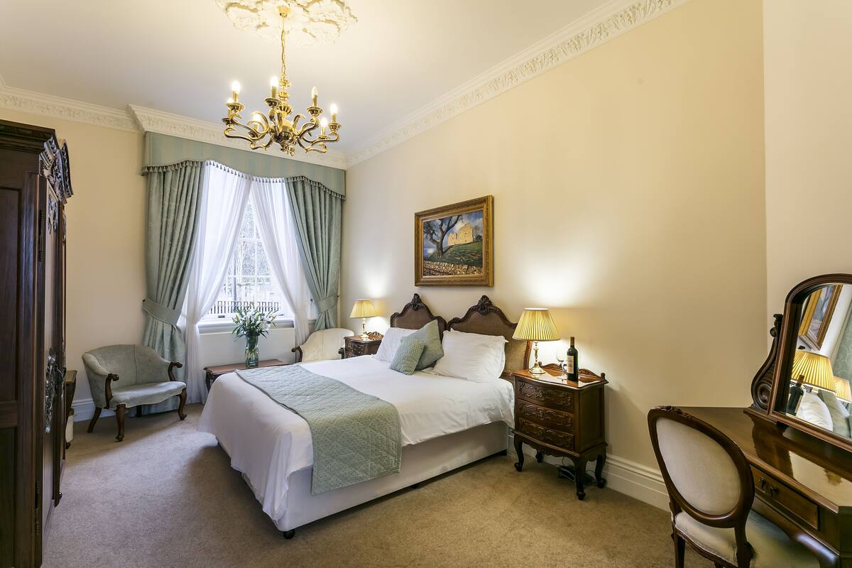 A bedroom with a large bed, a chair, a desk, a dresser, a chandelier, and a painting on the wall.