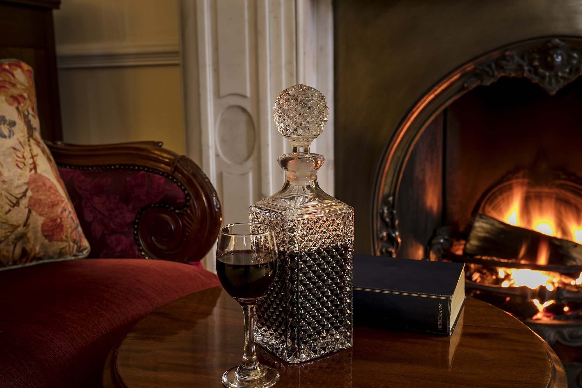 A decanter, a wine glass, and a book on a table in a room with a fireplace.