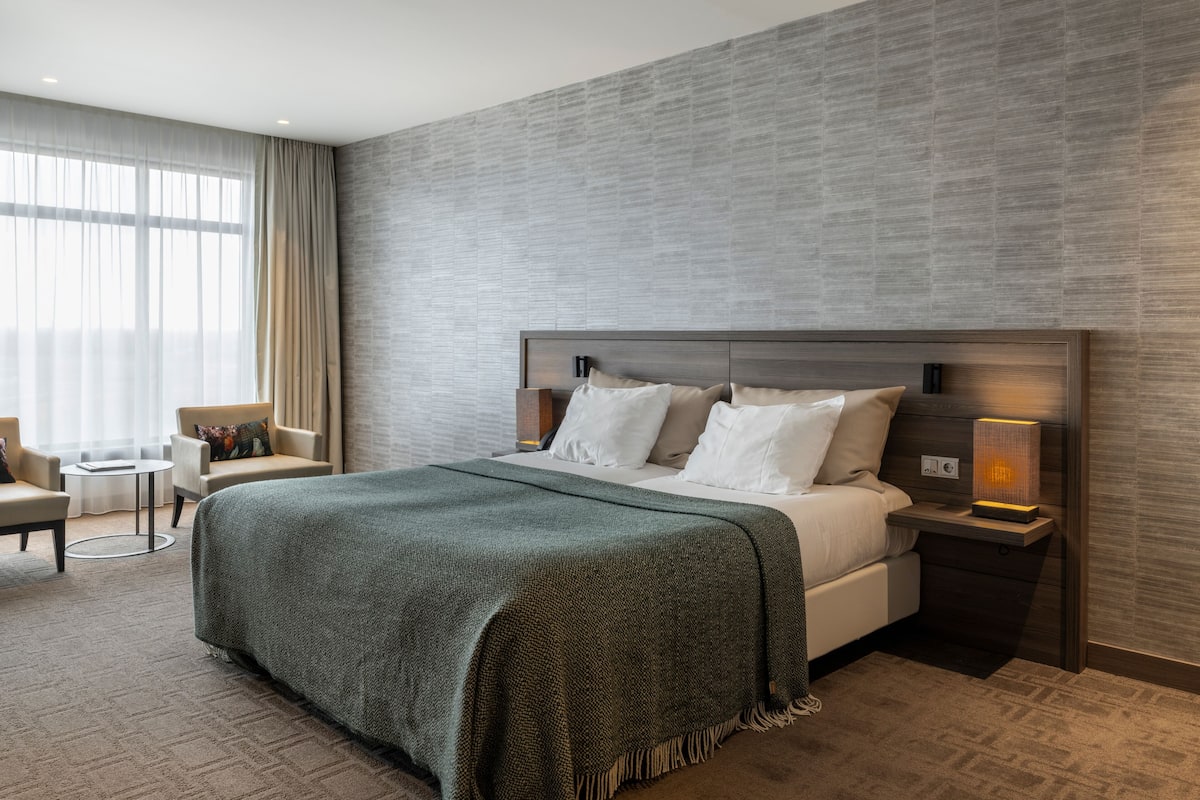 A modern hotel room with a large bed, a bedside table with a lamp, a small round table, and a chair.