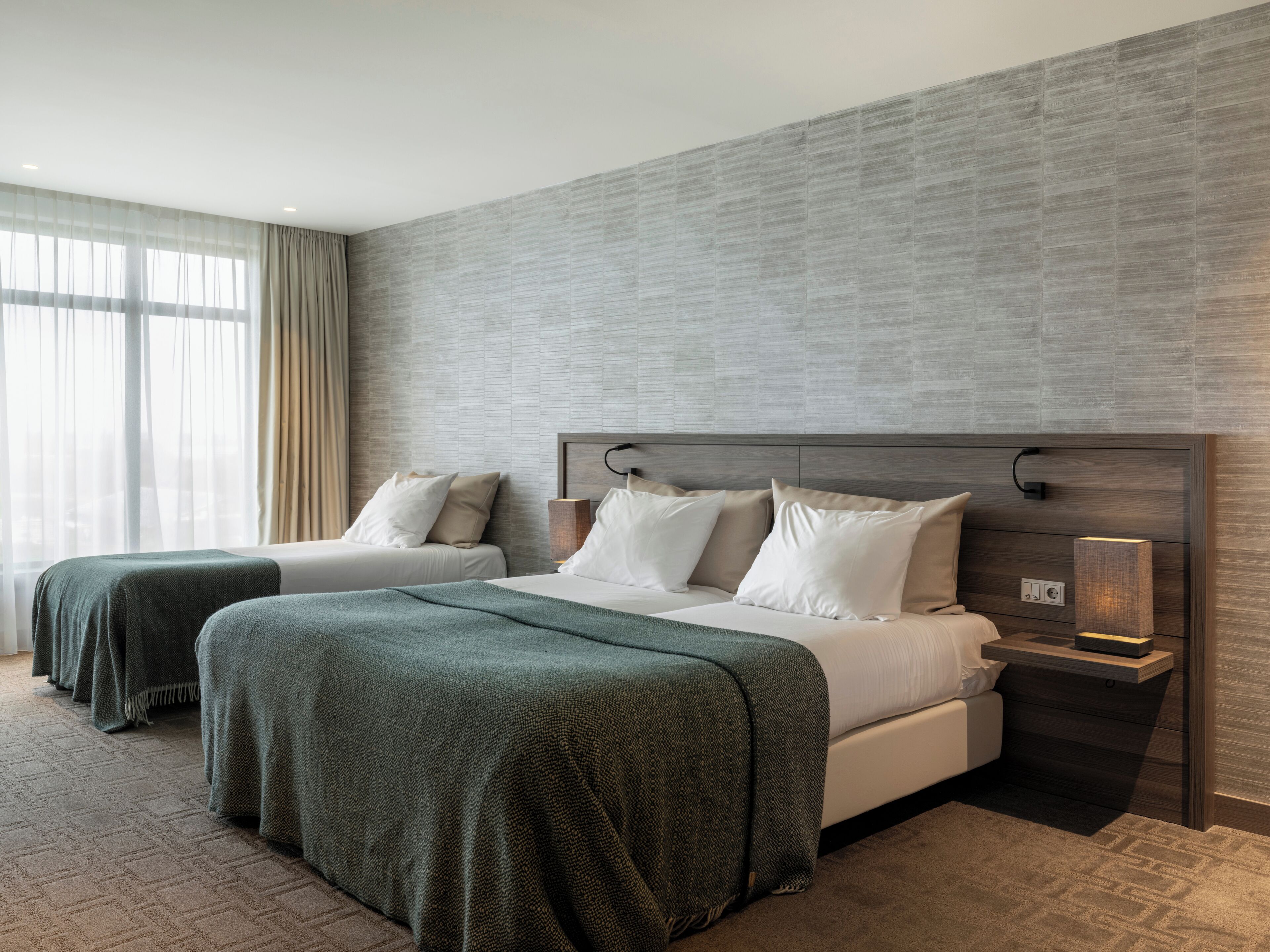 A hotel room with two beds, a large window with curtains, a textured wall, and a bedside table with a lamp.