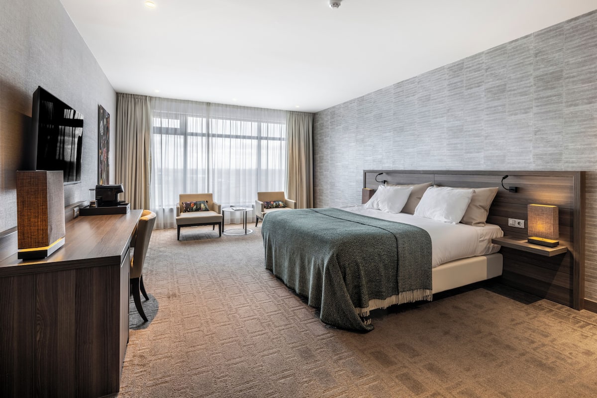 A modern hotel room with a large bed, a desk with a flat-screen TV, two armchairs, a coffee machine, and a large window with sheer curtains.