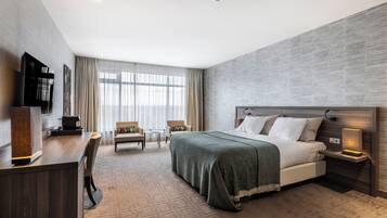 Een moderne hotelkamer met een groot bed, een bureau met een flatscreen televisie, twee fauteuils, een koffiezetapparaat en een groot raam met dunne gordijnen.