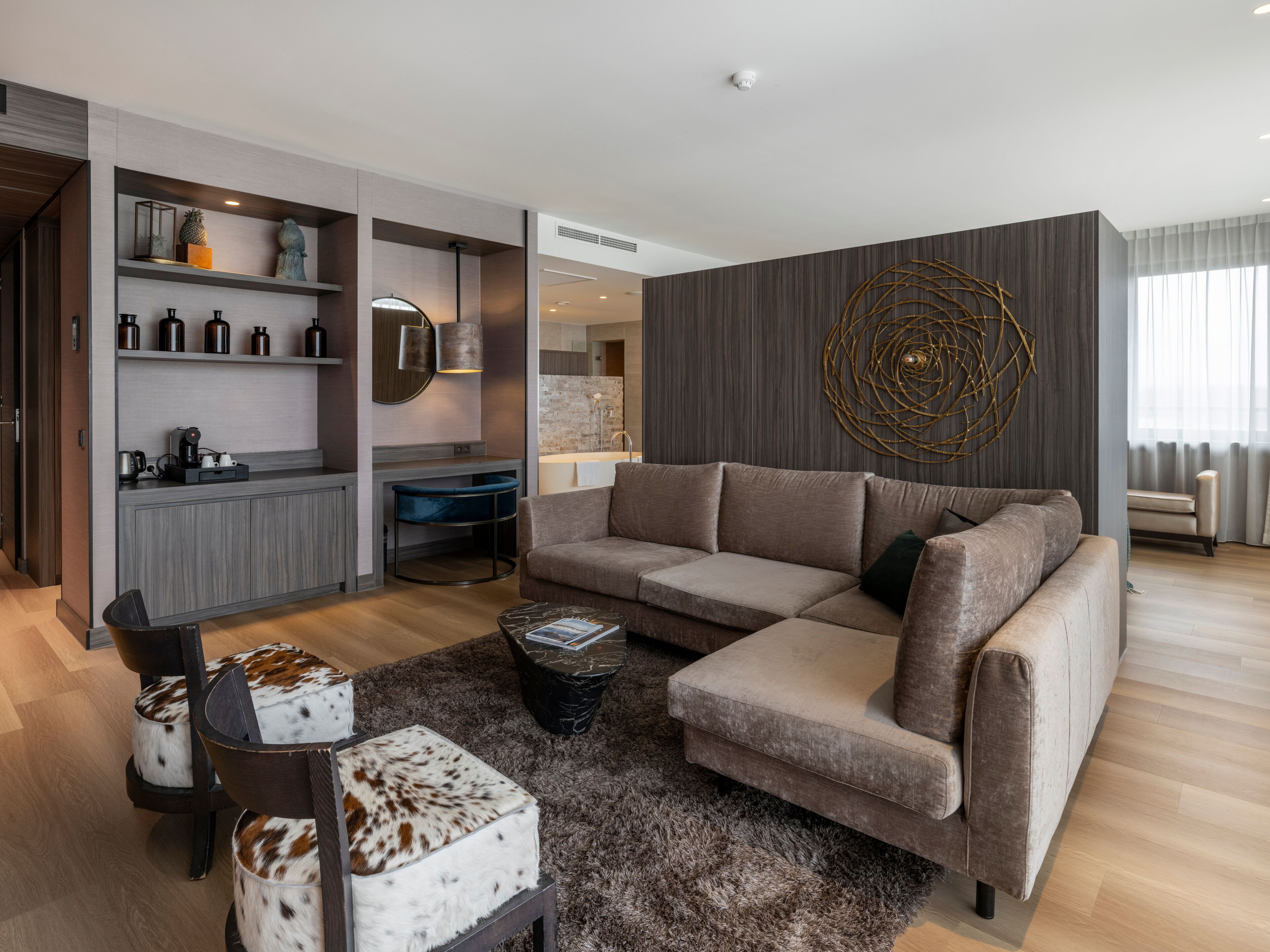 Una sala de estar moderna con un sofá modular, una mesa de centro y una alfombra mullida. Se ve un área de cocina con una encimera y un lavamanos al fondo.
