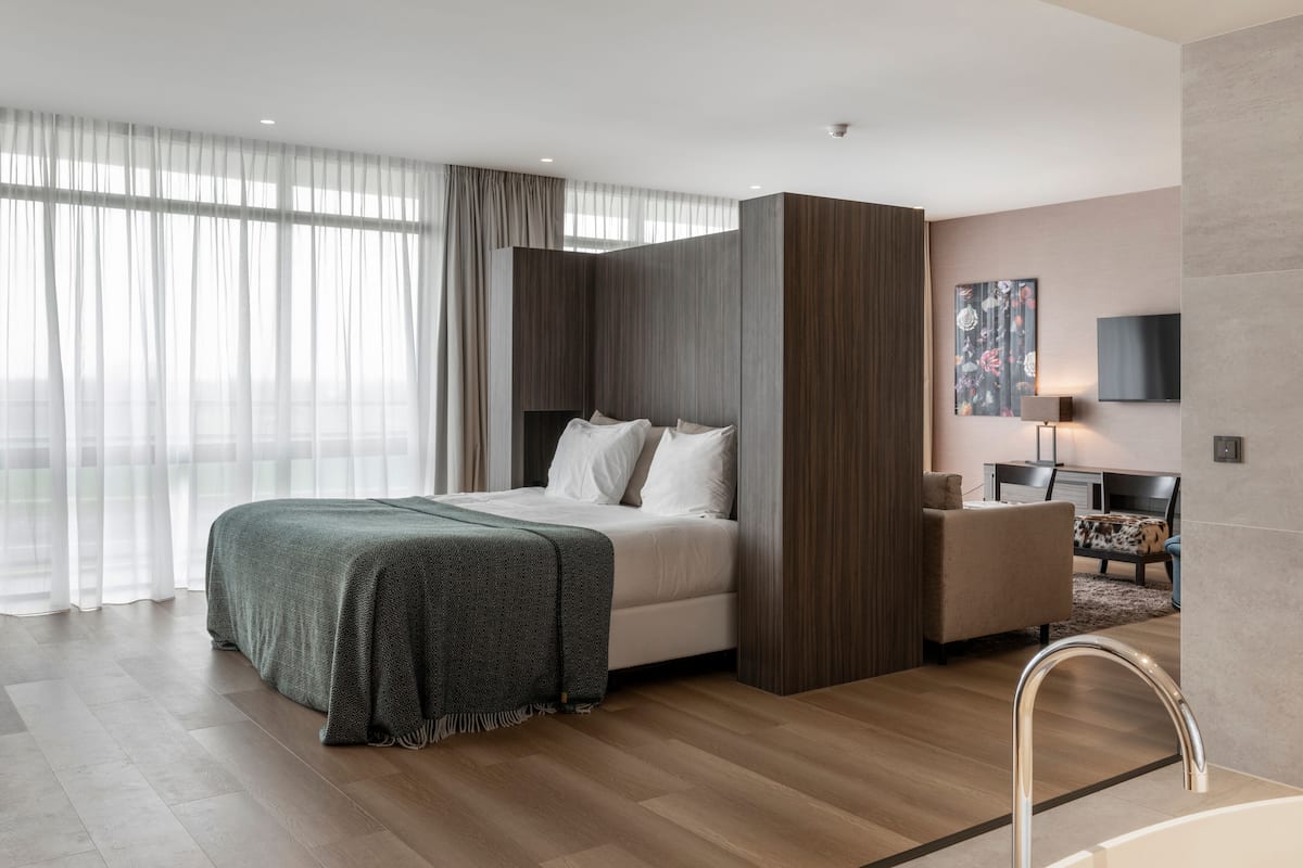 A modern hotel room with a large bed, a desk, a chair, and a bathroom with a sink and mirror.