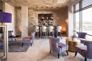 A modern bar area with a geometric ceiling, bar stools, and a large window with a view of parked cars. - Van der Valk Hotel Zwolle (Zwolle)