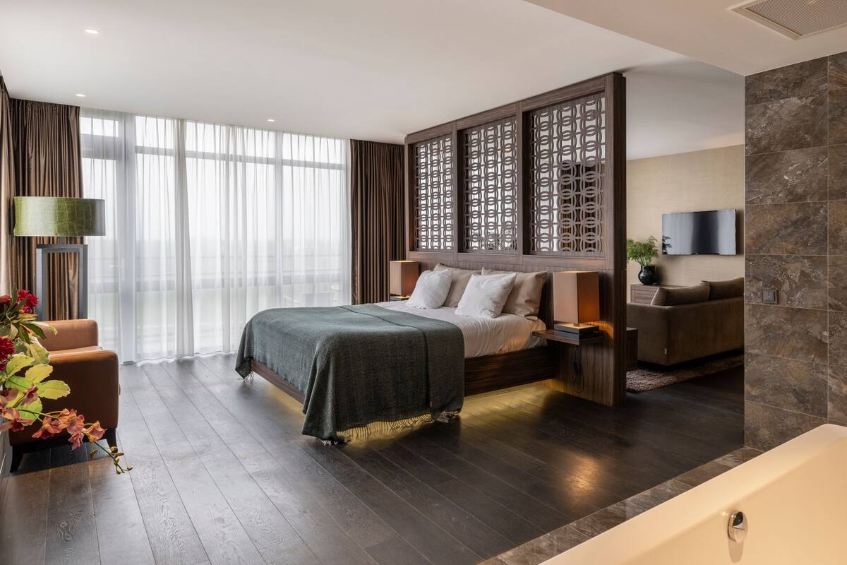 A modern hotel room with a large bed, a seating area, a TV, and a stone accent wall.
