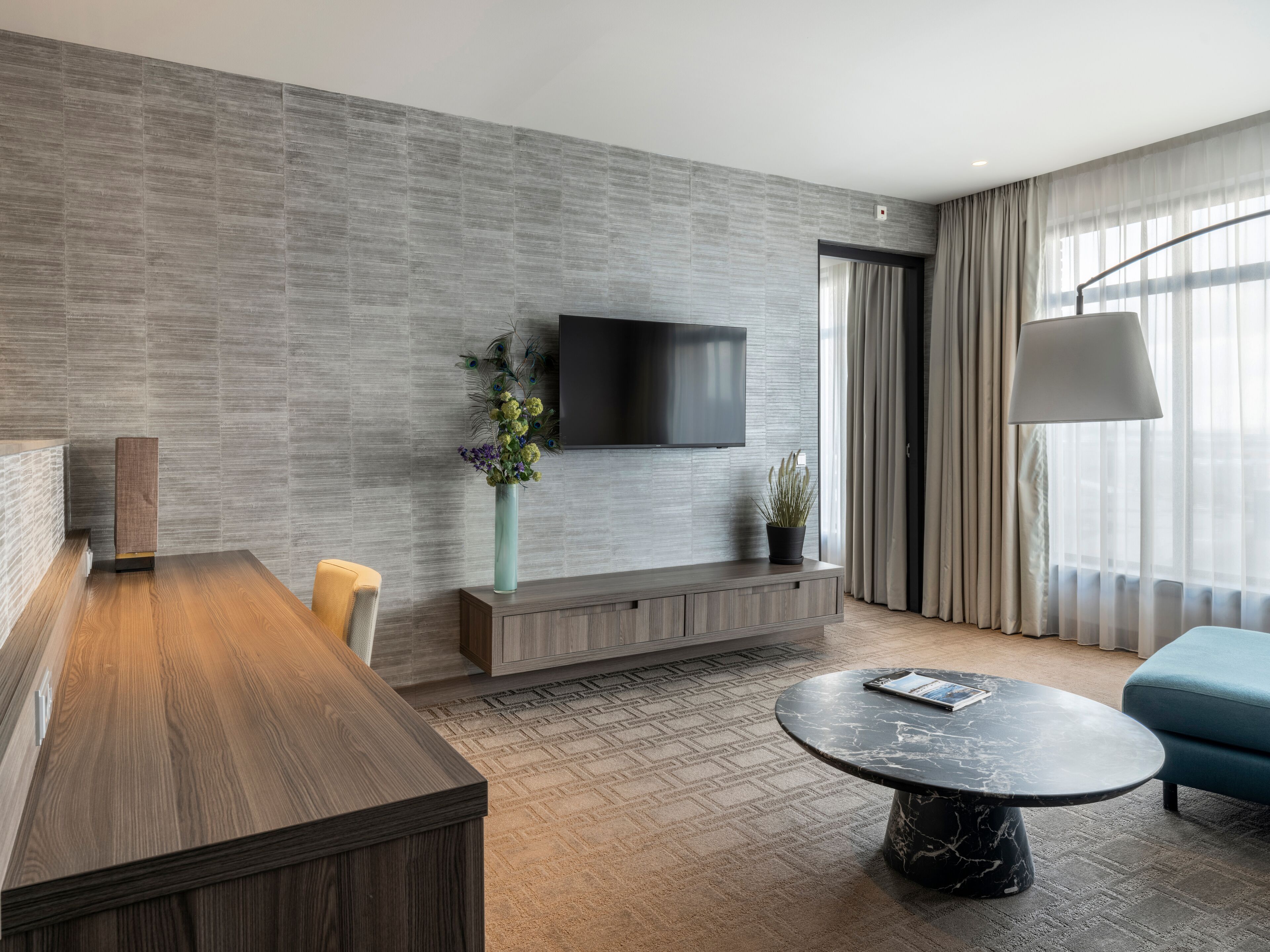 Een moderne hotelkamer met een houten eettafel, een ronde marmeren salontafel, een flatscreen televisie aan een gestructureerde wand, een potplant en een blauwe bank.