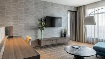 Een moderne hotelkamer met een houten eettafel, een ronde marmeren salontafel, een flatscreen televisie aan een gestructureerde wand, een potplant en een blauwe bank.