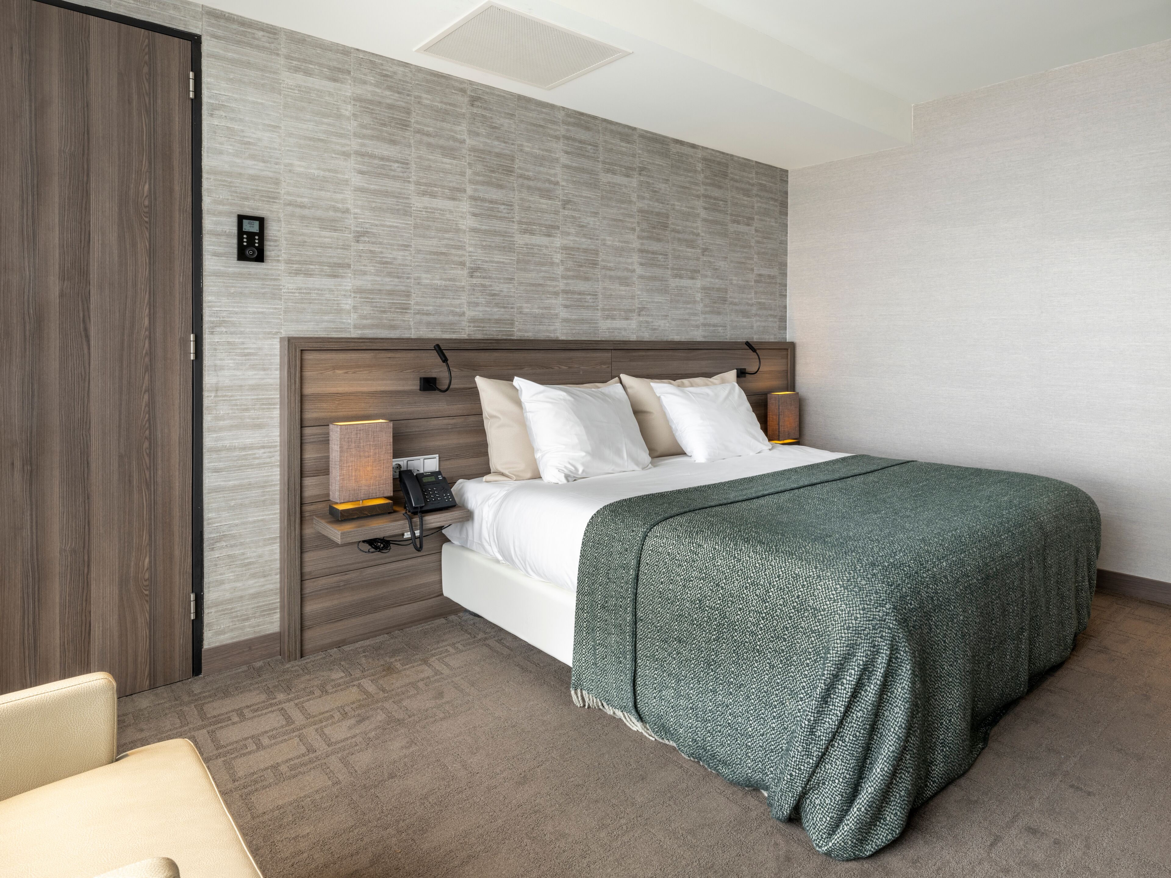 A hotel room with a wooden headboard, a bed with white linens, a green throw blanket, a bedside table with a lamp, and a wall-mounted phone.