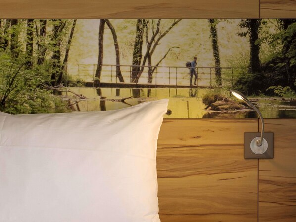Quarto com piso de madeira, travesseiro branco e luminária fixada na parede.
