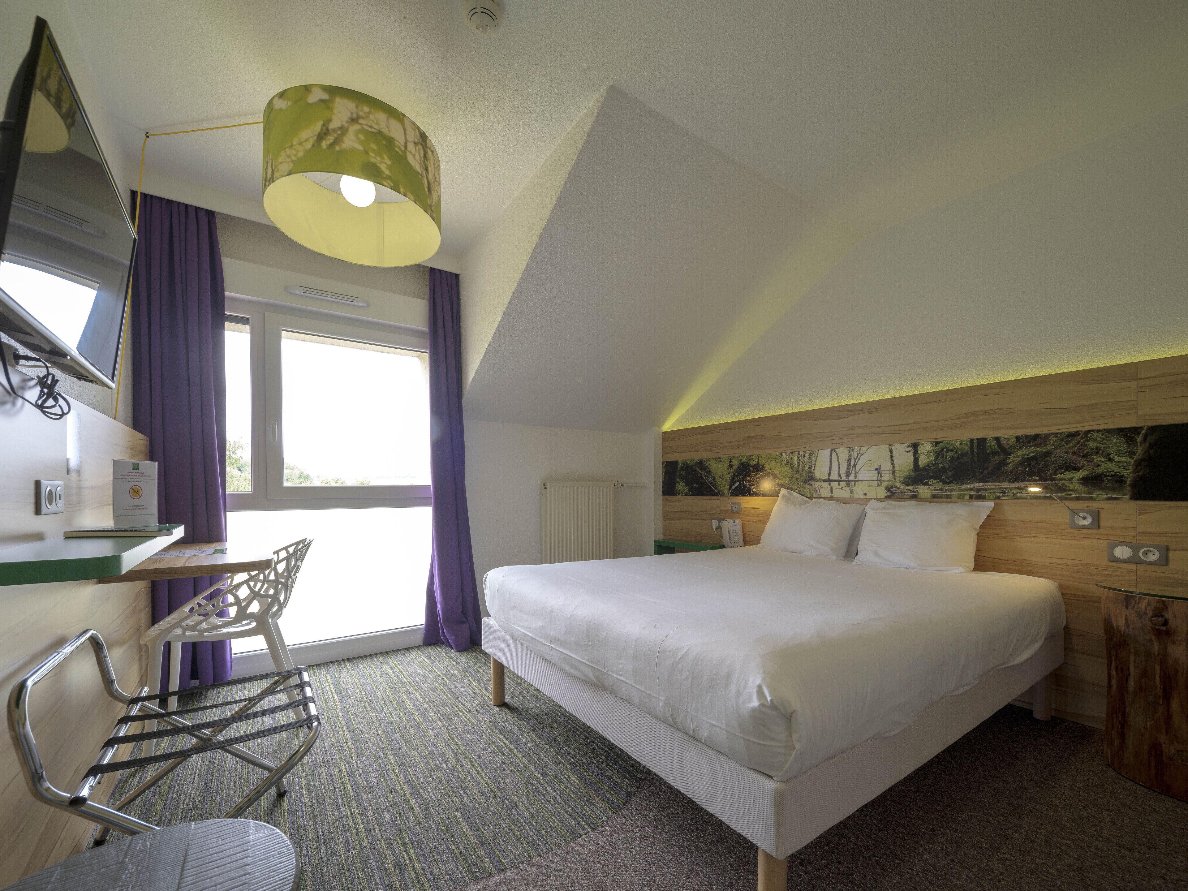 Ein Hotelzimmer mit Bett, Schreibtisch, Stuhl, einem Fenster mit Vorhängen und einer Wandlampe.
