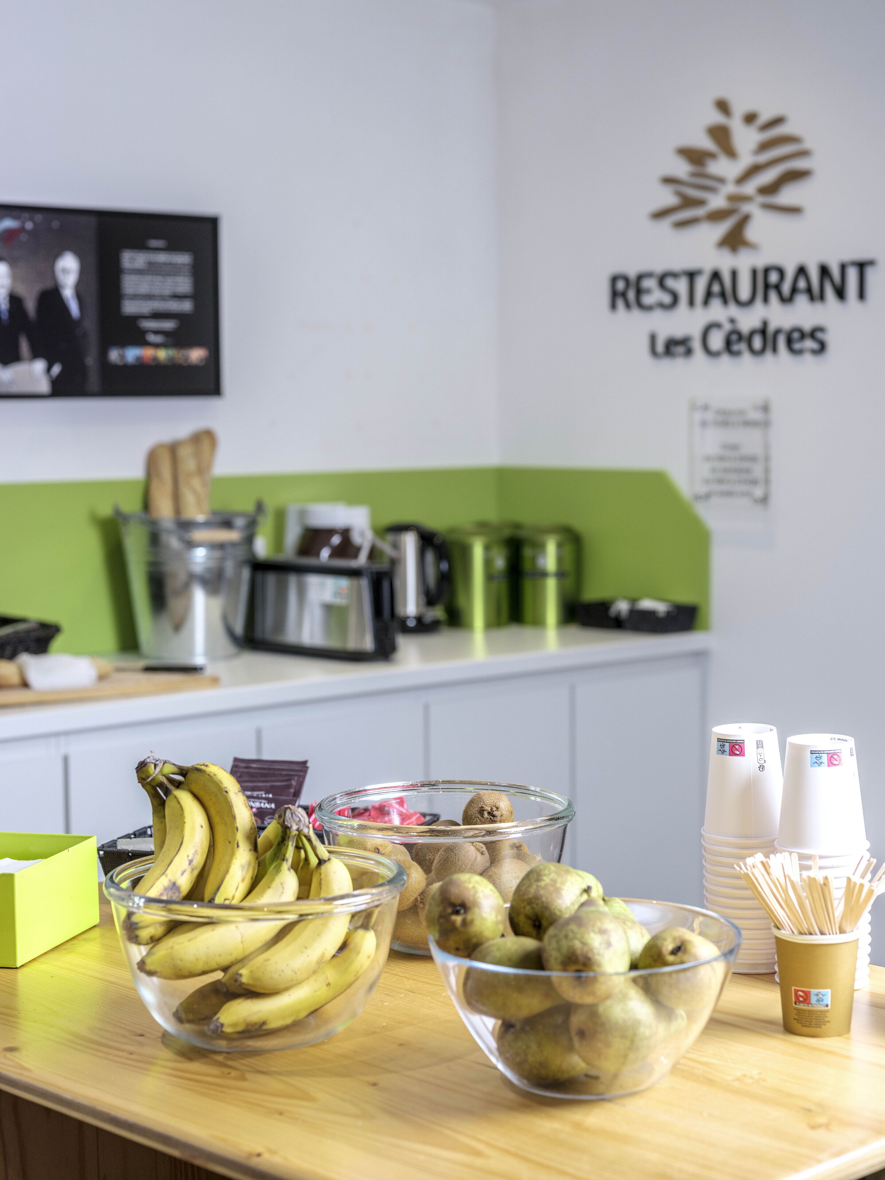 A kitchen counter with bowls of fruit, a coffee machine, and a sign for 'RESTAURANT Les Cendres'.
