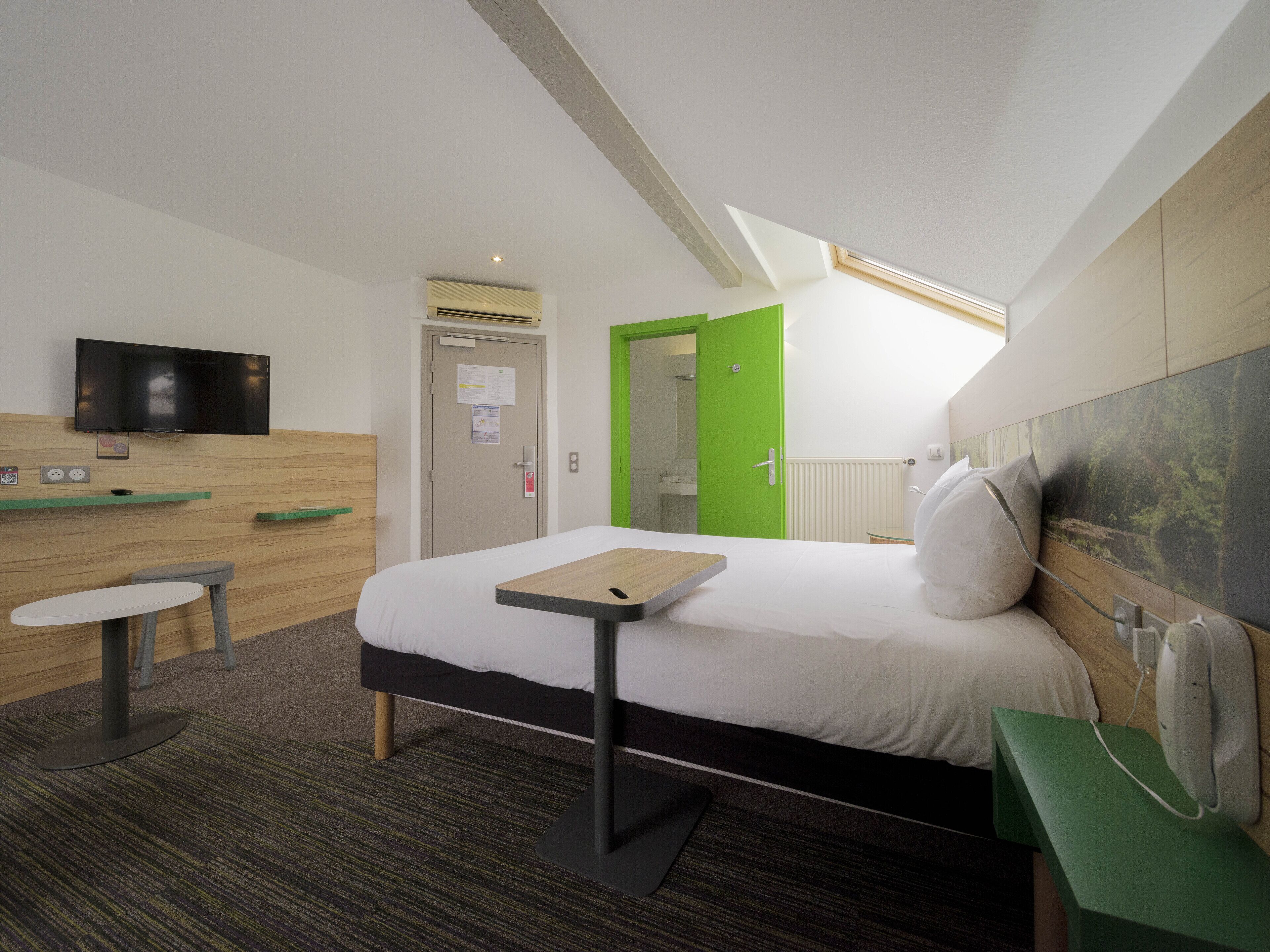 A hotel room with a bed, bedside table, a flat-screen TV mounted on the wall, a green door, and a window with a view of the sky.