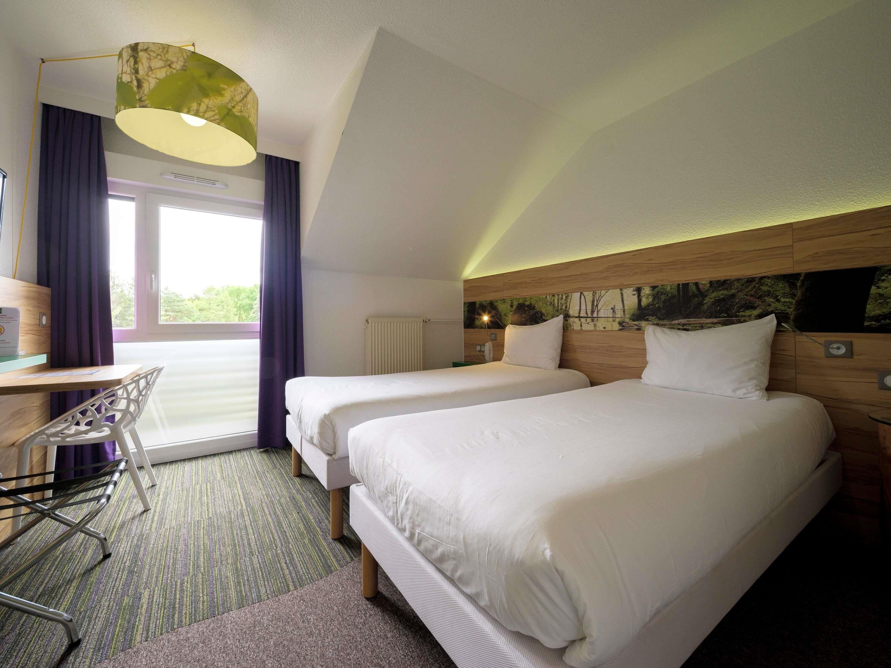 A hotel room with two beds, a desk, a chair, and a large window with curtains.