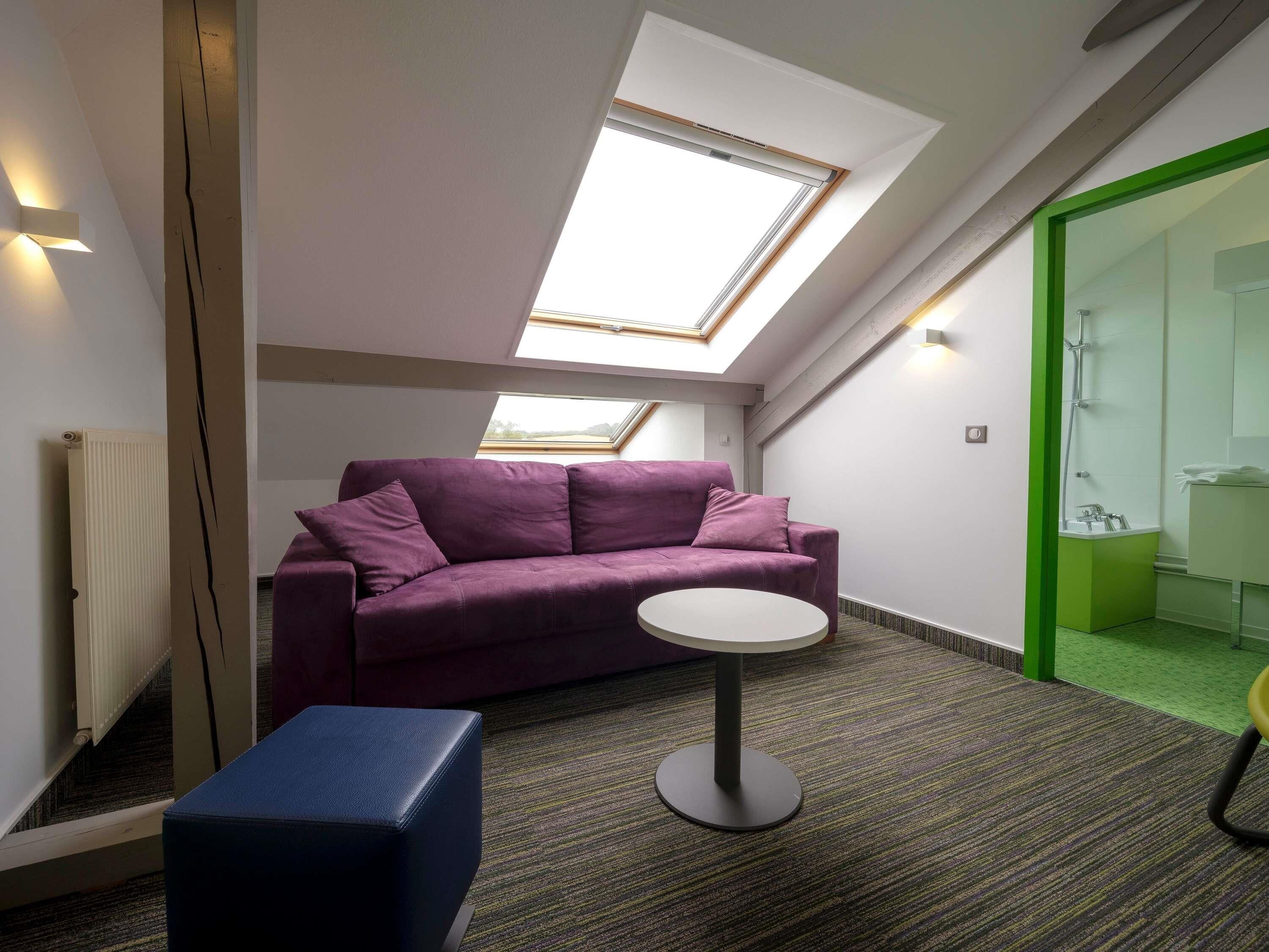 A room with a purple sofa, a small round table, a blue ottoman, a skylight, and a green-framed shower area.