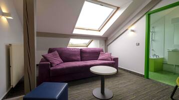 A room with a purple sofa, a small round table, a blue ottoman, a skylight, and a green-framed shower area.