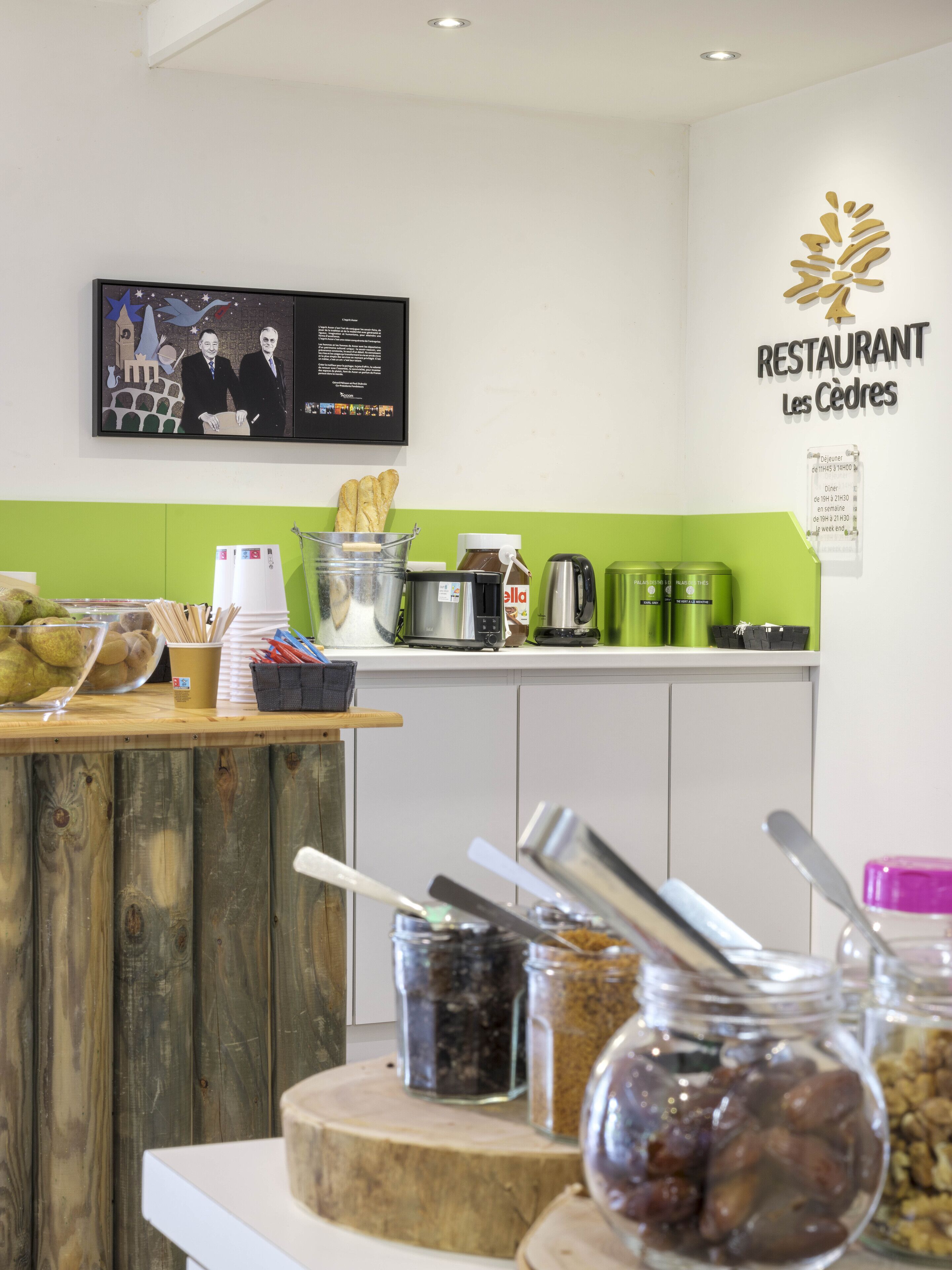 Una cocina de restaurante con una pared verde, un televisor mostrando un menú y diversos utensilios de cocina e ingredientes sobre el mostrador.