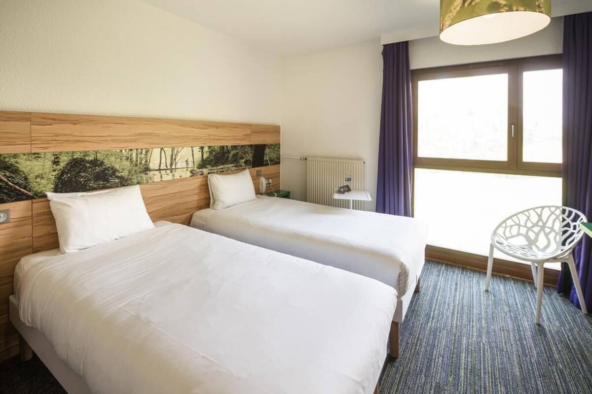 Ein Hotelzimmer mit zwei Betten, einem Fenster mit Vorhängen, einem Heizkörper, einem Stuhl und einer Lampe.