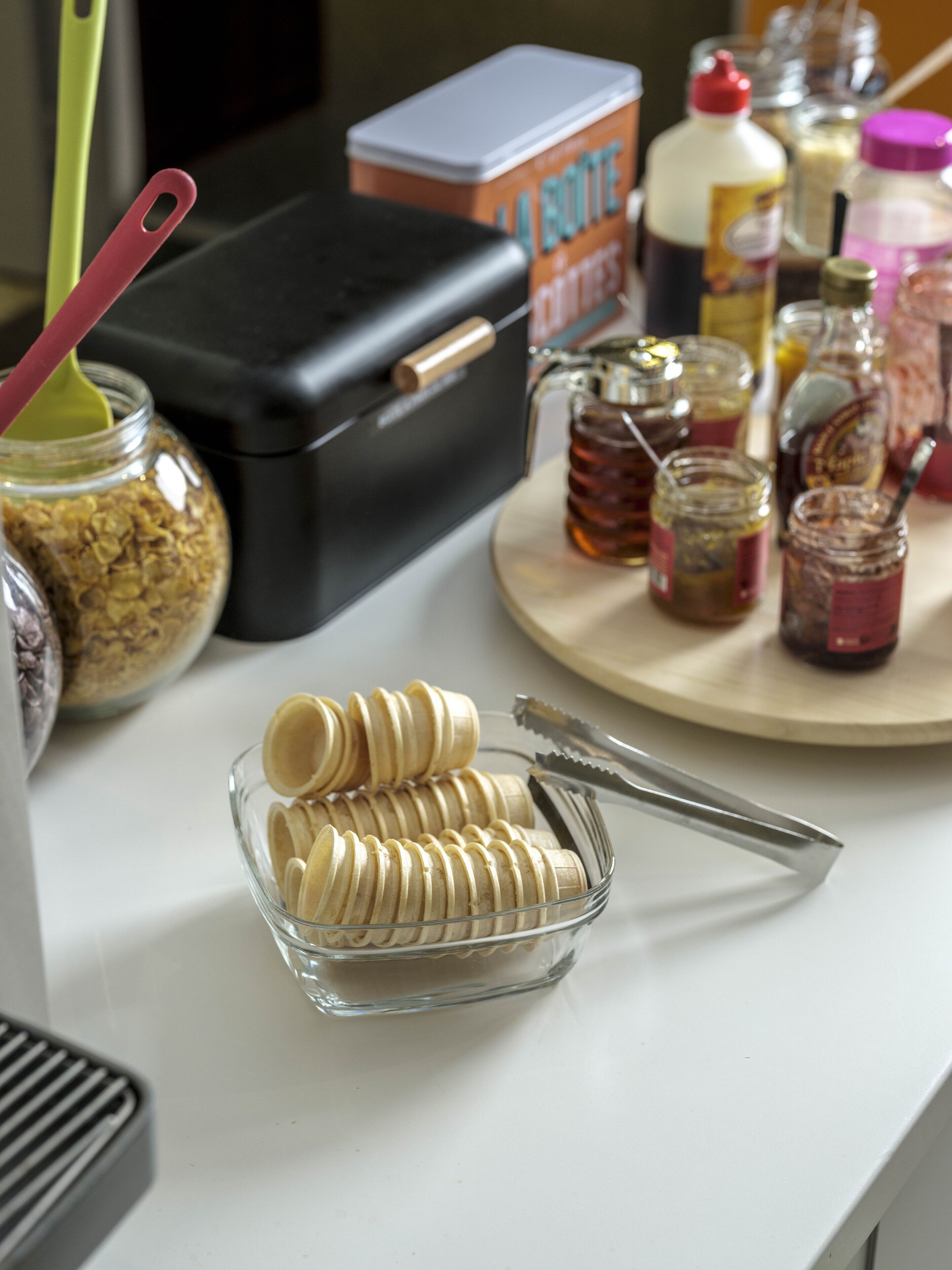 Una encimera de cocina con frascos de condimentos, una caja de azúcar y un bol de vidrio con rebanadas de pan.