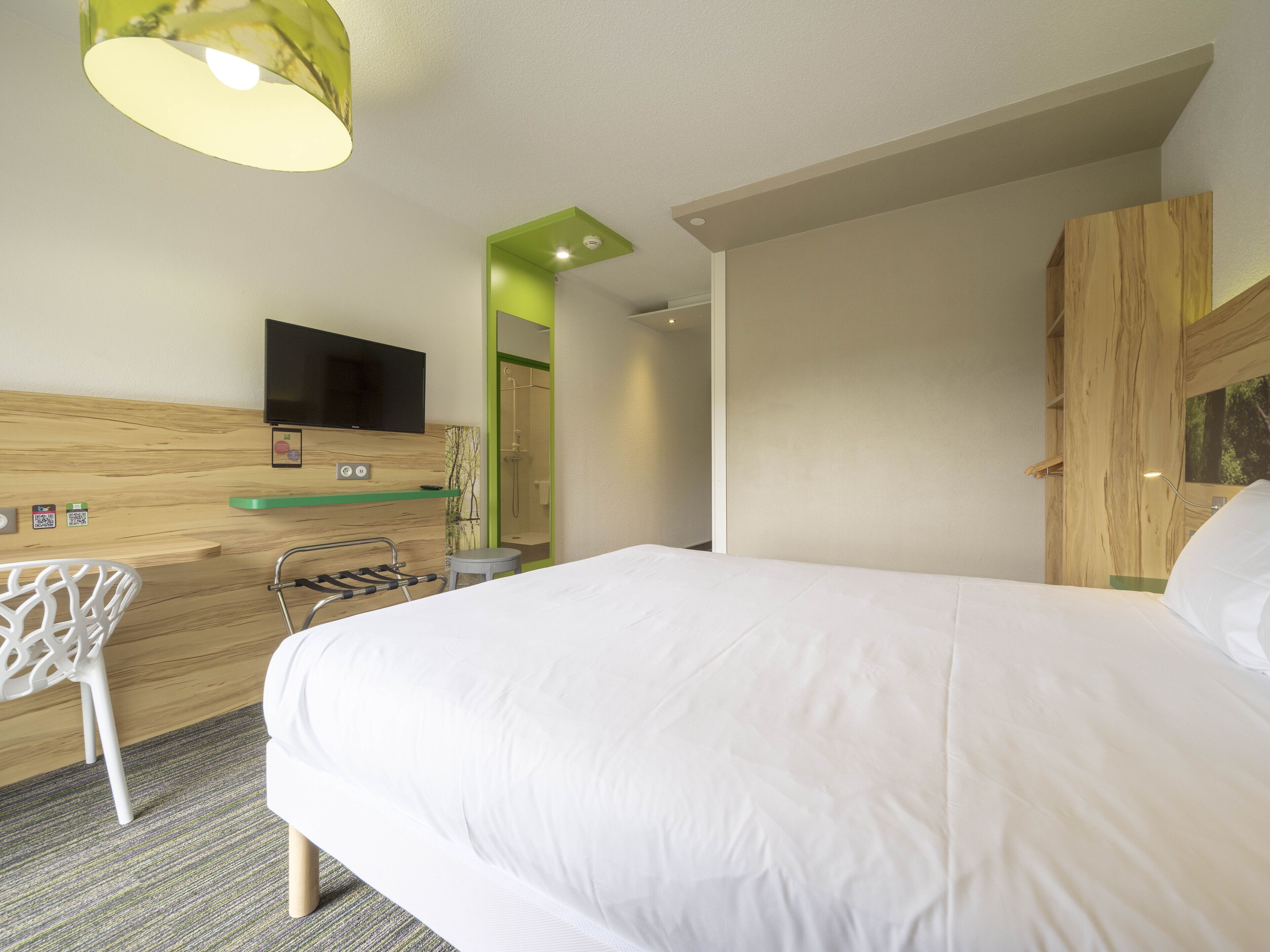 A hotel room with a bed, a desk with a chair, a TV, and a shower area.