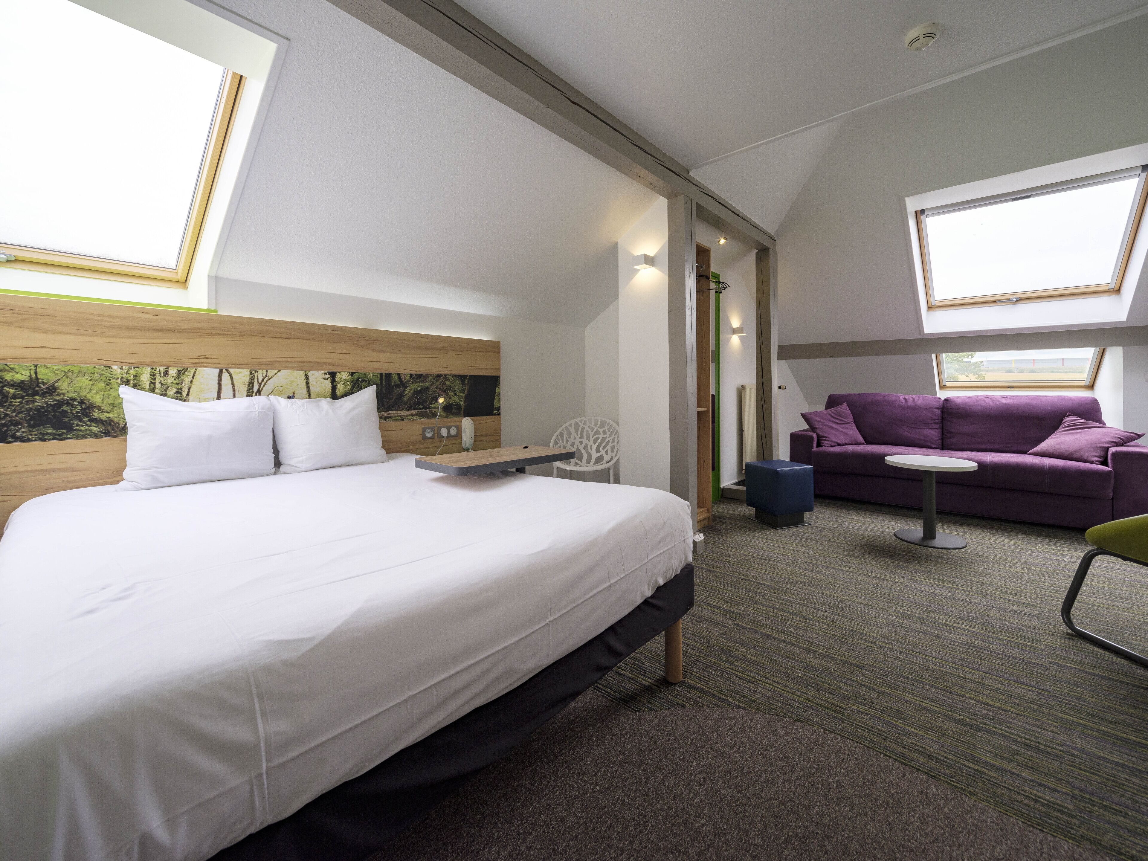 A modern hotel room with a large bed, a purple sofa, a small table, and a chair.