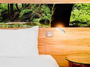 Un dormitorio con cabecera de madera, almohada blanca, mesita de noche y vistas a un bosque y al agua.